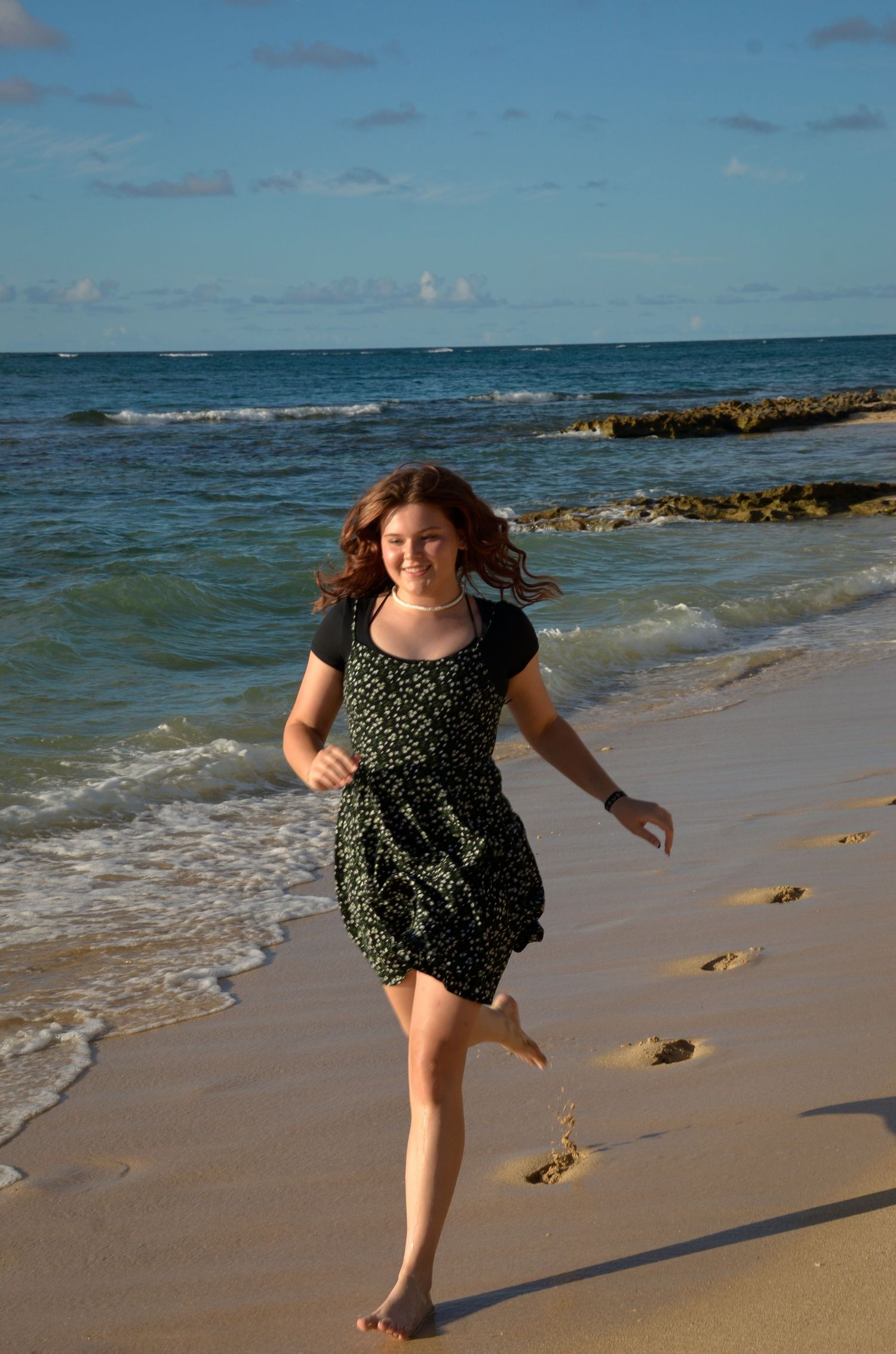 Person in floral dress runs joyfully along beach shoreline at sunset.