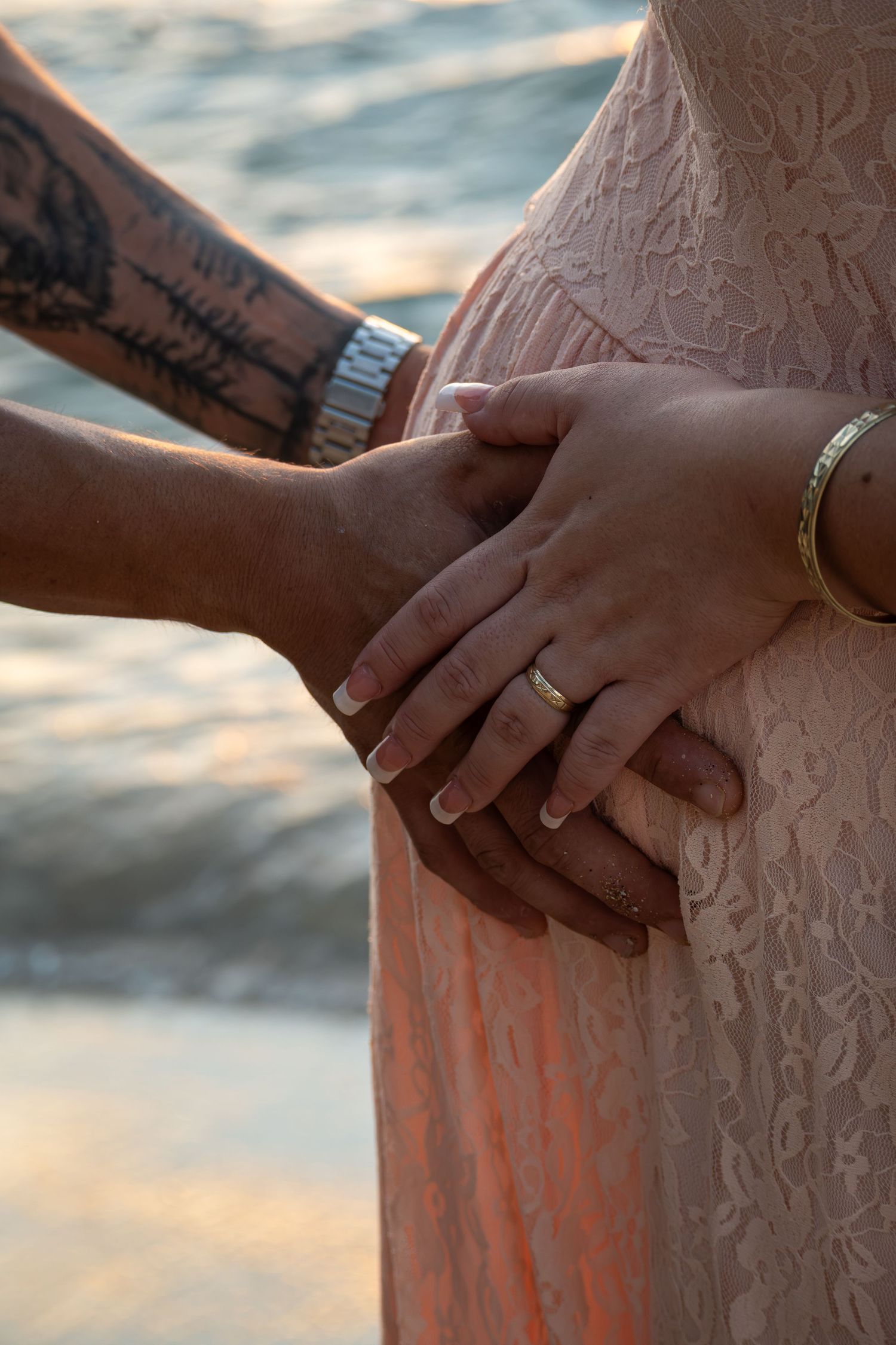 Couple's hands resting on pregnant belly, man with tattoo and watch, woman with ring and bracelet, near water.