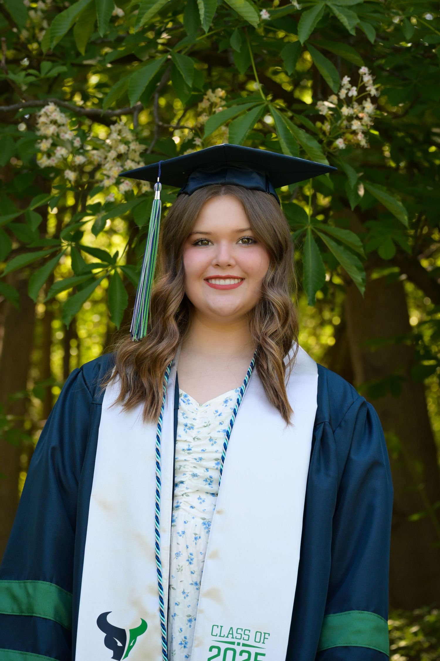 Graduate in cap and gown with green and white stole poses outdoors against leafy background.
