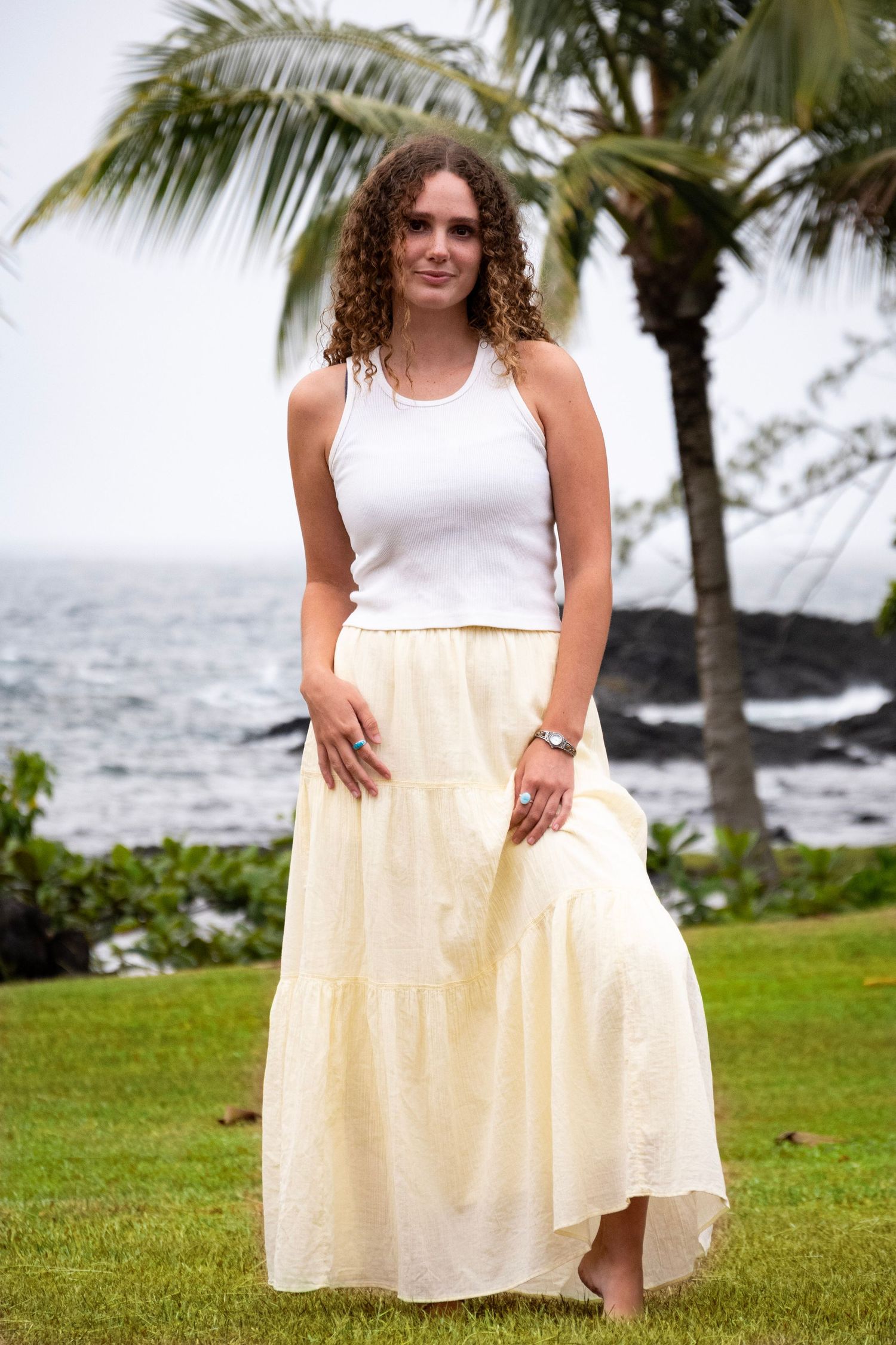 Person in a white tank top and flowing cream maxi skirt poses on a grassy oceanfront with palm trees in Hawaii.