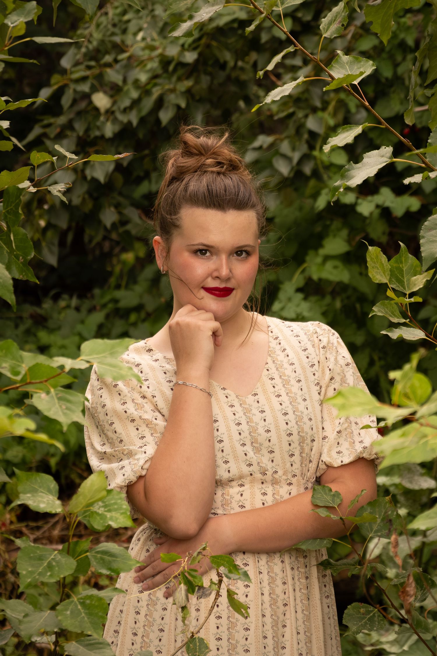 Young woman in floral dress with red lips poses confidently among lush green ivy leaves outdoors.