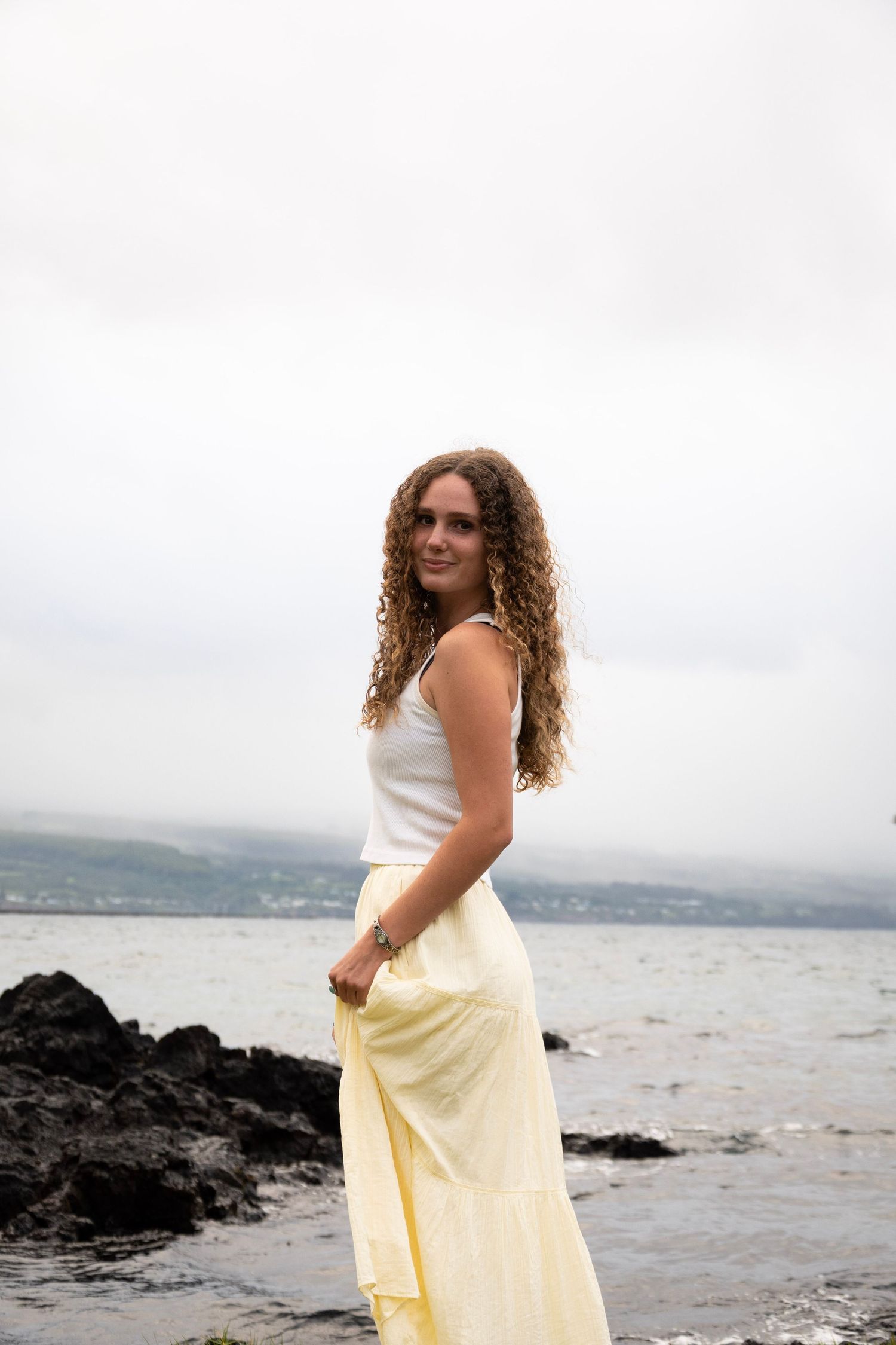 A figure in a white top and yellow skirt poses gracefully along a rocky beach shoreline with mountains in the distance.