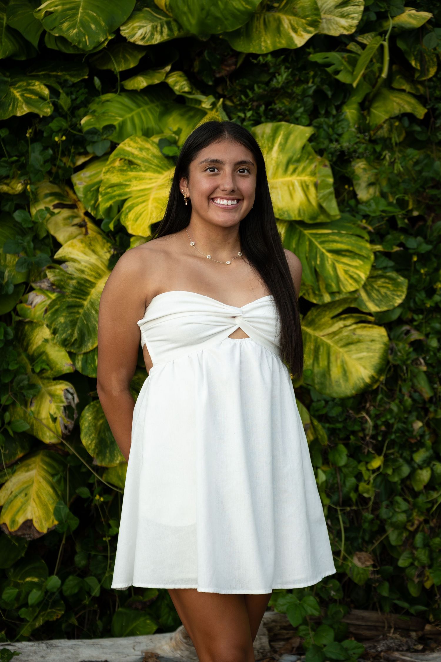 A woman in a white off-shoulder top poses against a backdrop of lush green tropical foliage.
