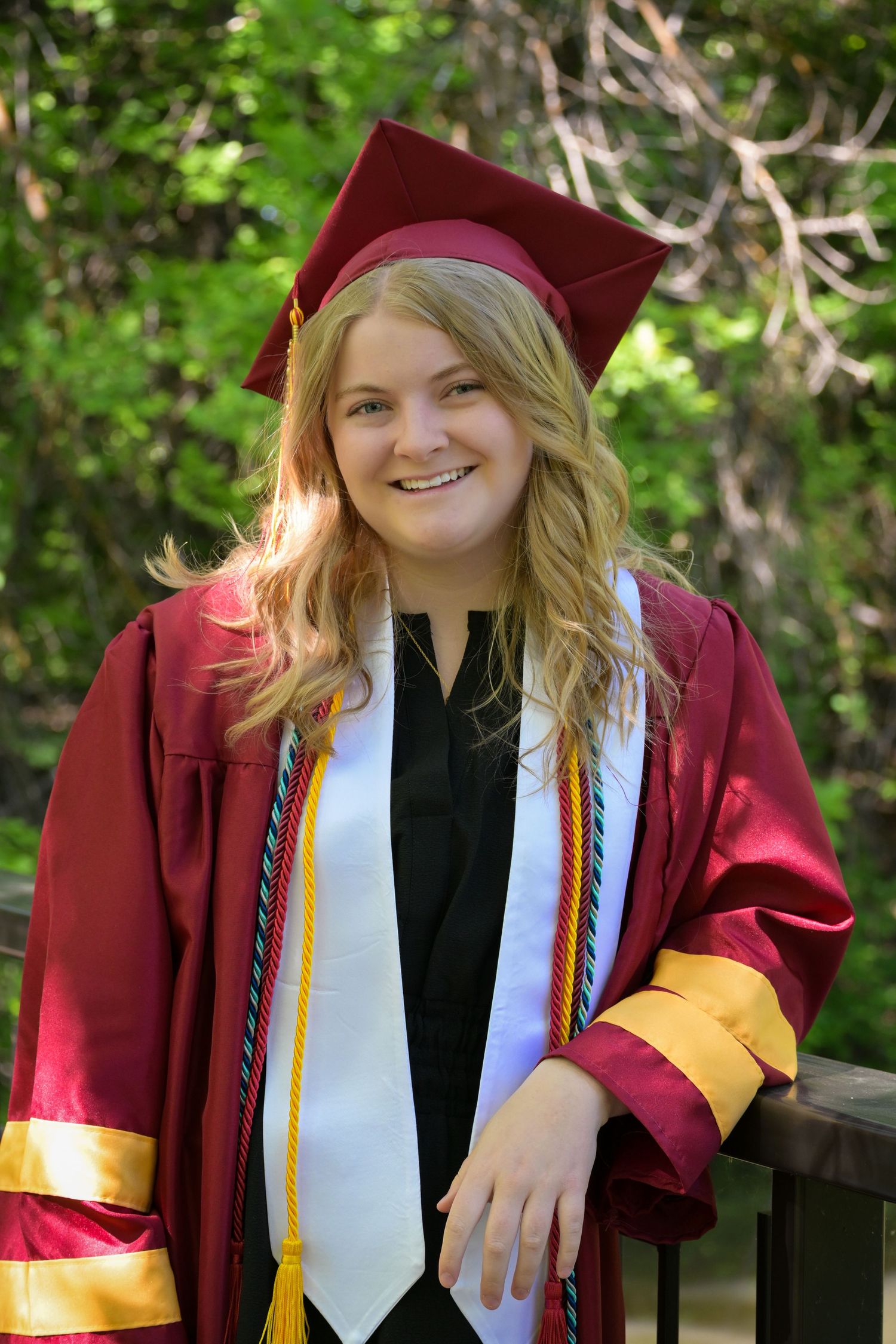 Graduate wearing maroon and gold academic regalia poses outdoors against green foliage background.
