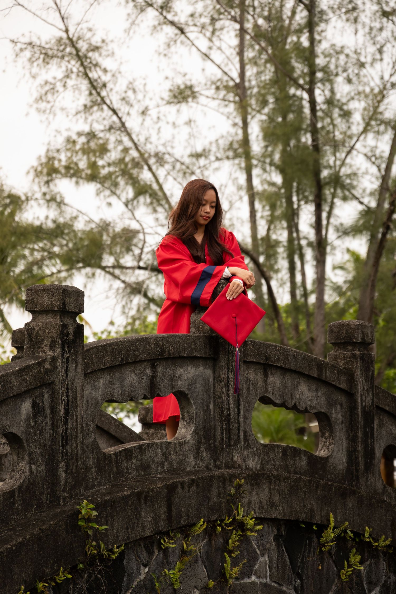 Young woman in red graduation gown and cap standing on ornate stone bridge surrounded by trees.