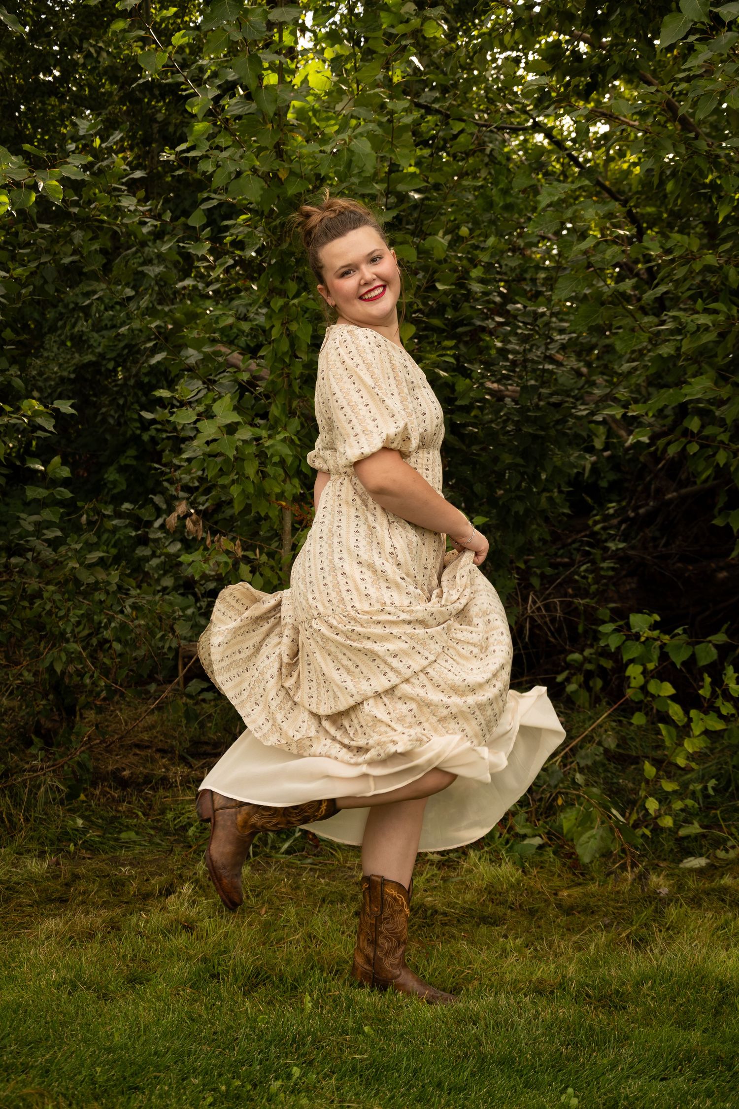 Woman in floral prairie dress and cowboy boots twirling outdoors in lush green garden.