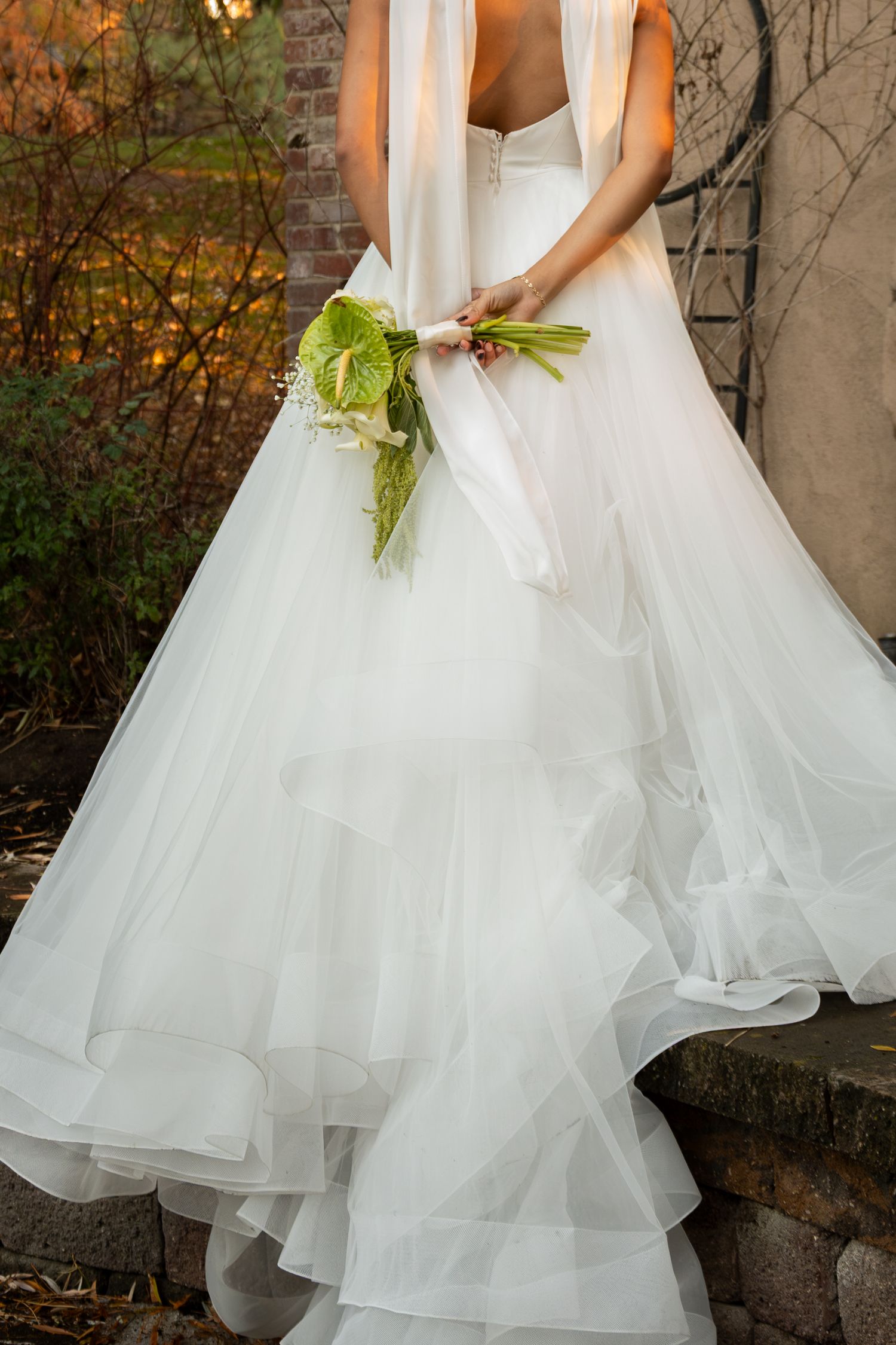 Bride in layered tulle wedding gown holds a green and white bouquet while standing on stone steps outdoors.