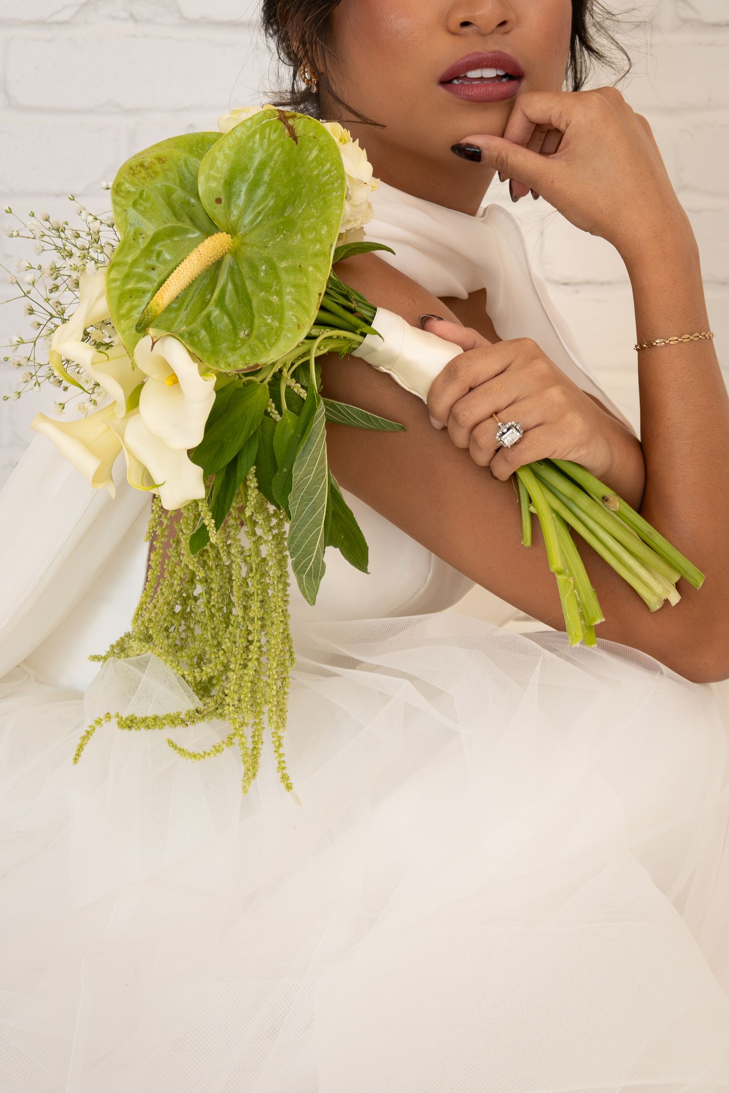 Bride in white gown holding green anthurium and calla lily bouquet with diamond ring visible.