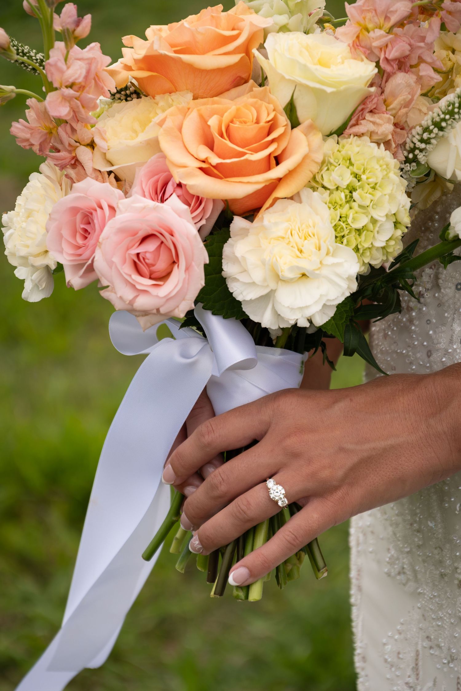 Bride holding a wedding bouquet of peach, pink, and white roses with a white ribbon, wearing a diamond engagement ring.