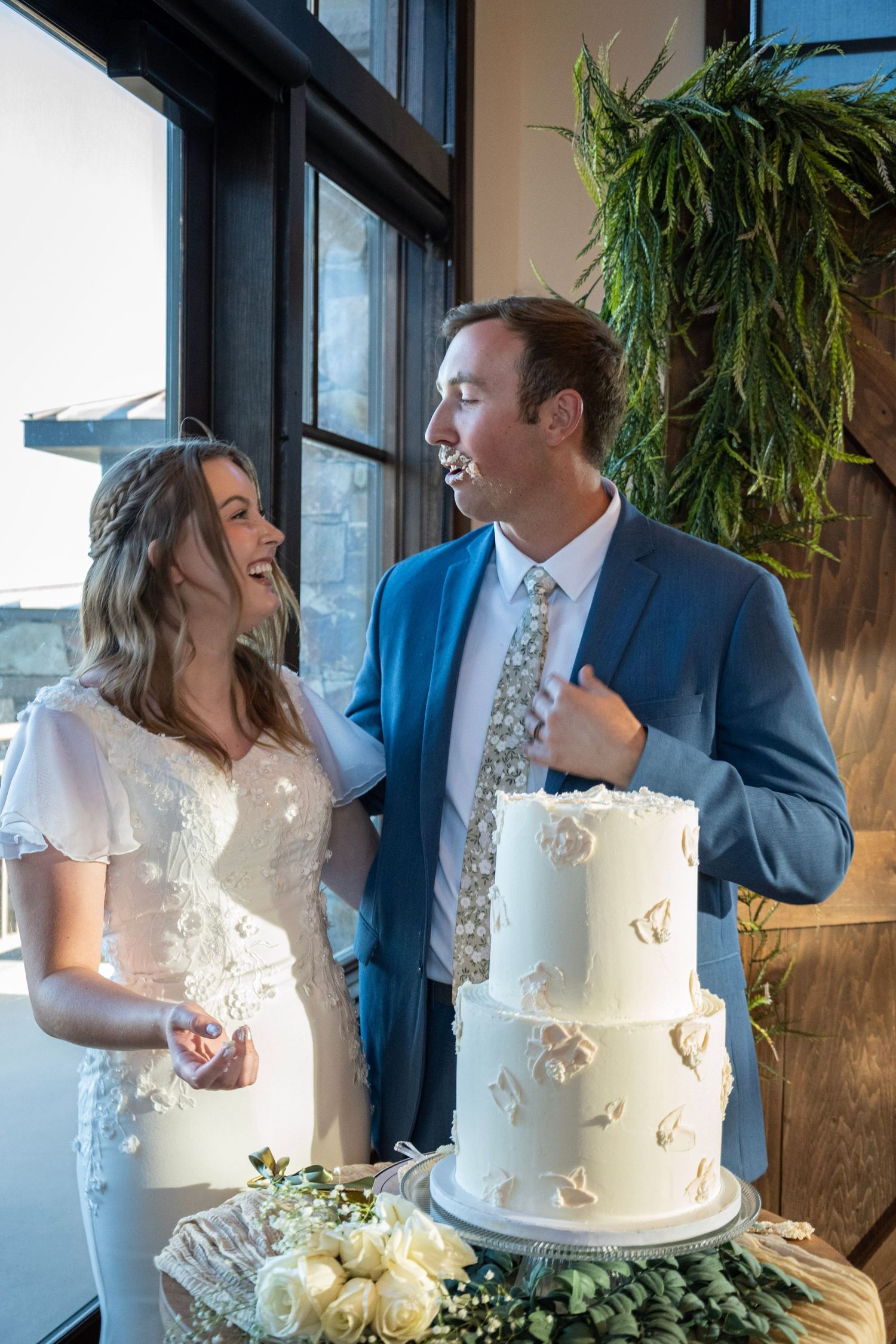 A couple shares laughter next to their elegant white two-tier wedding cake decorated with floral details.