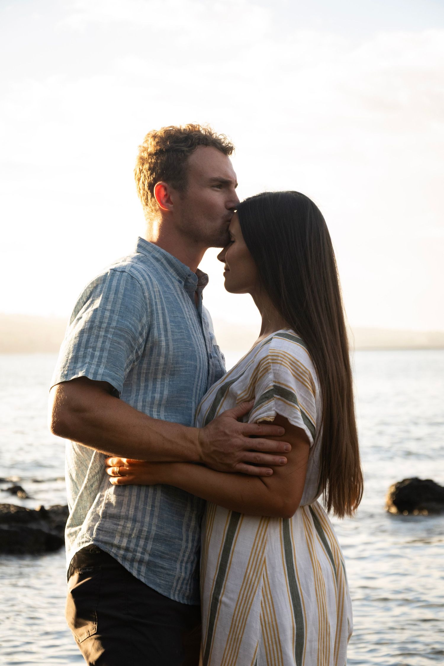 A romantic couple embraces on a rocky beach at sunset, with one person gently kissing the other's forehead.