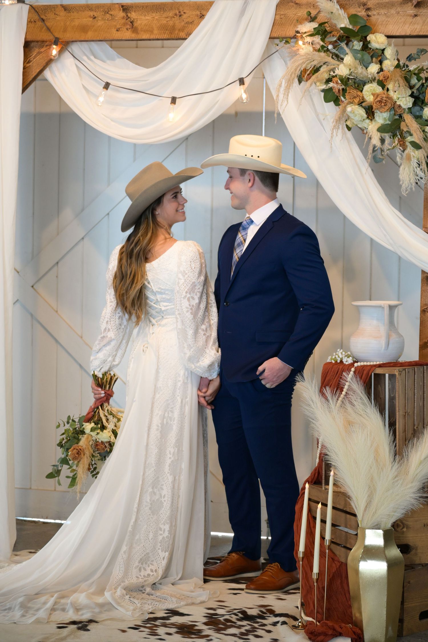 A Western-style wedding couple in cowboy hats pose under white draping and floral decor.