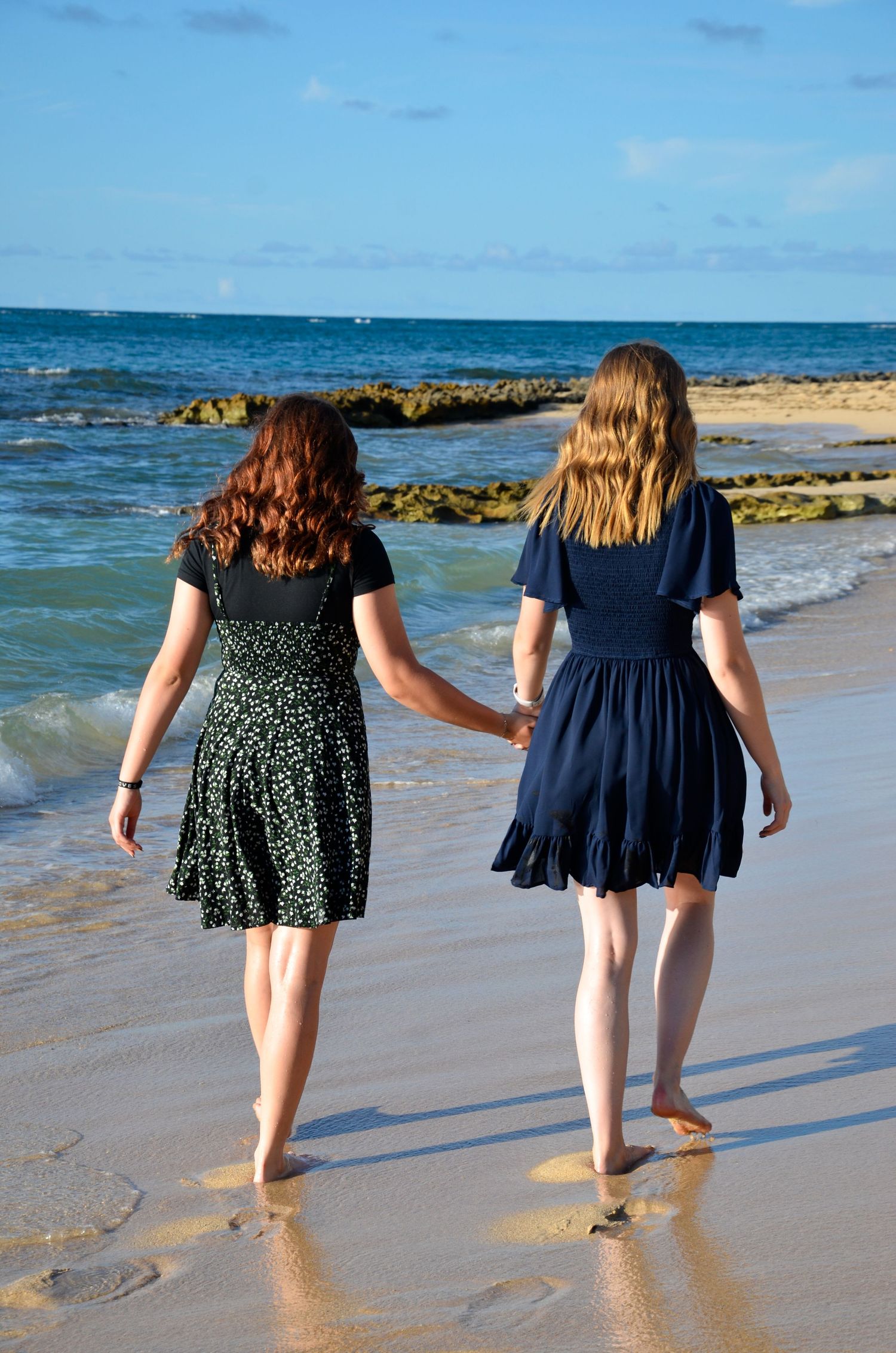 Two sisters in summer dresses walking along sandy beach shoreline.