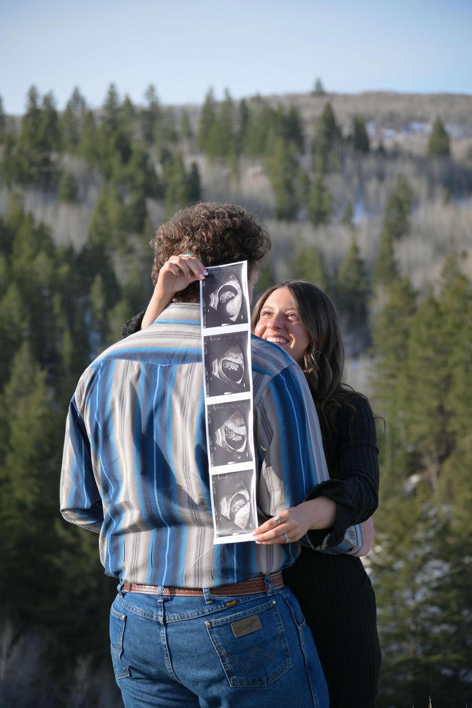 A couple sharing a happy moment while holding ultrasound photos against a forested mountain backdrop.