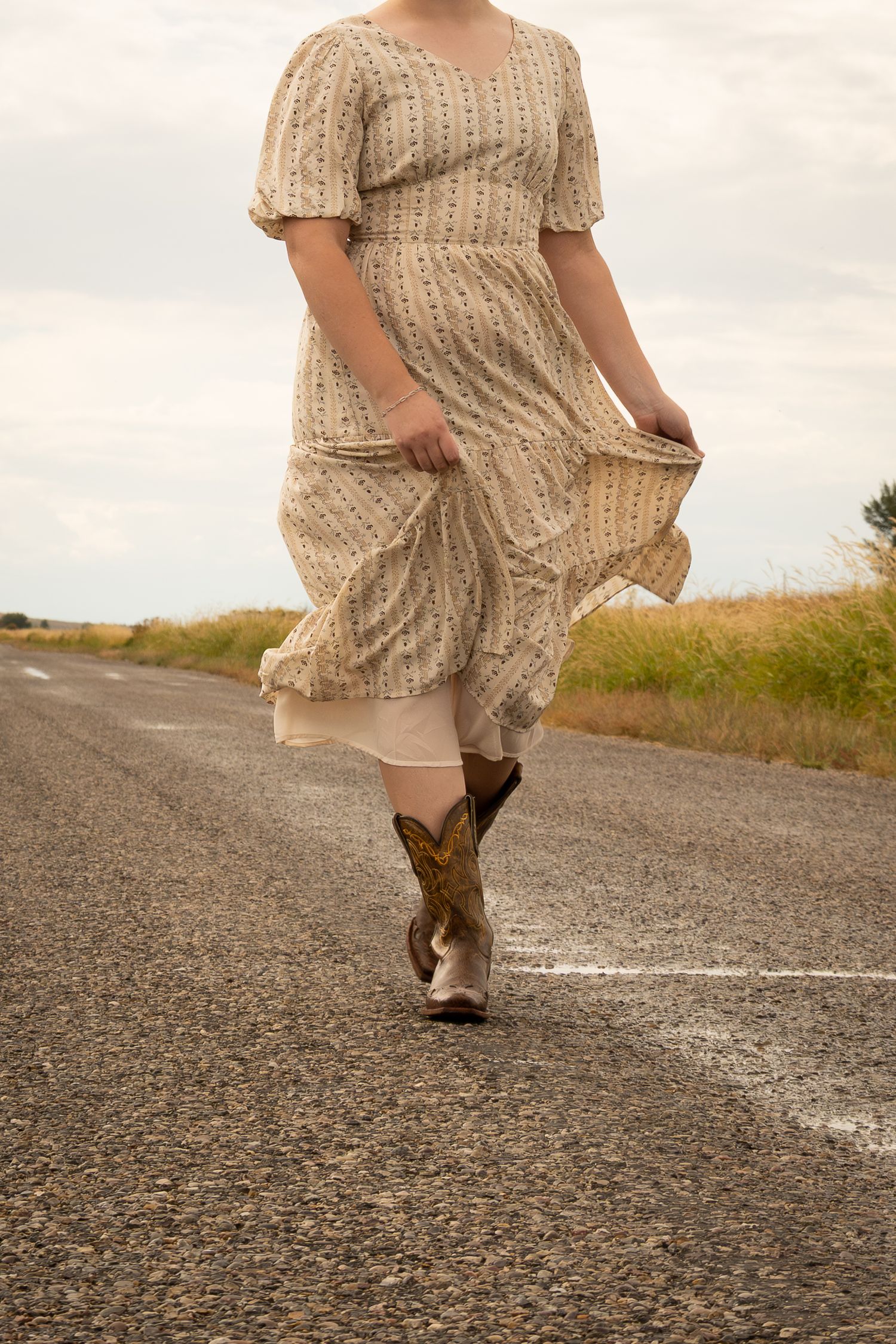 Woman in floral prairie dress and cowboy boots walking on a rural road, lifting her skirt in the breeze.
