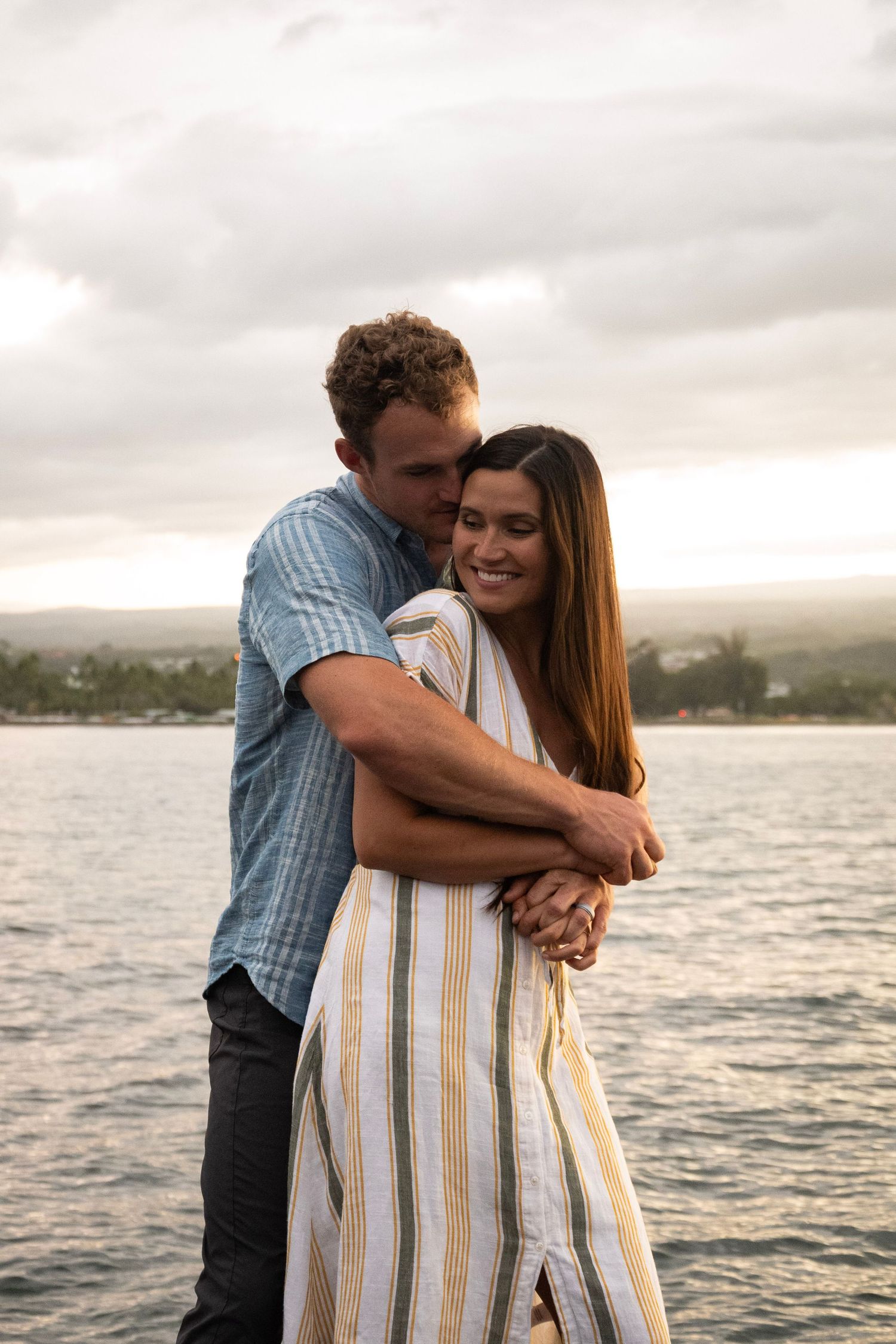 A couple shares an intimate embrace by a serene waterfront at dusk.