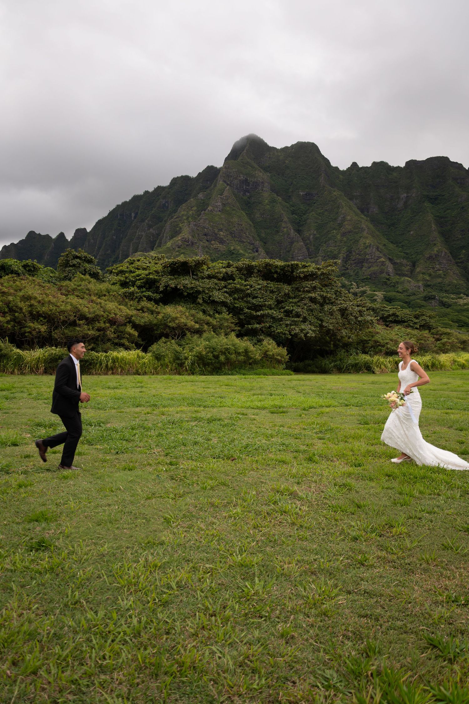 Bride and groom walking toward each other in a green meadow with dramatic misty mountain peaks in Hawaii.