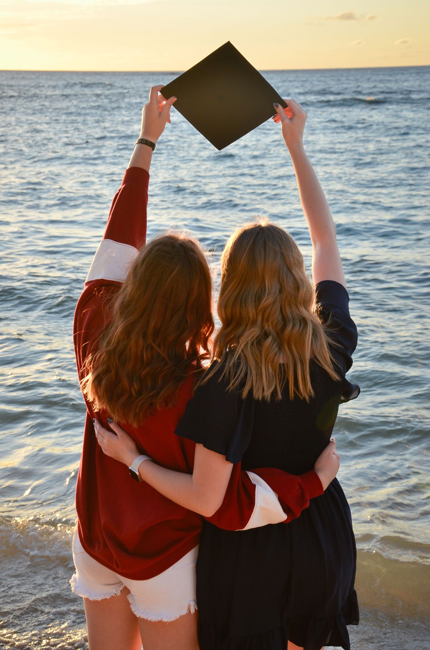 Two sisters holding graduation cap against sunset ocean view.