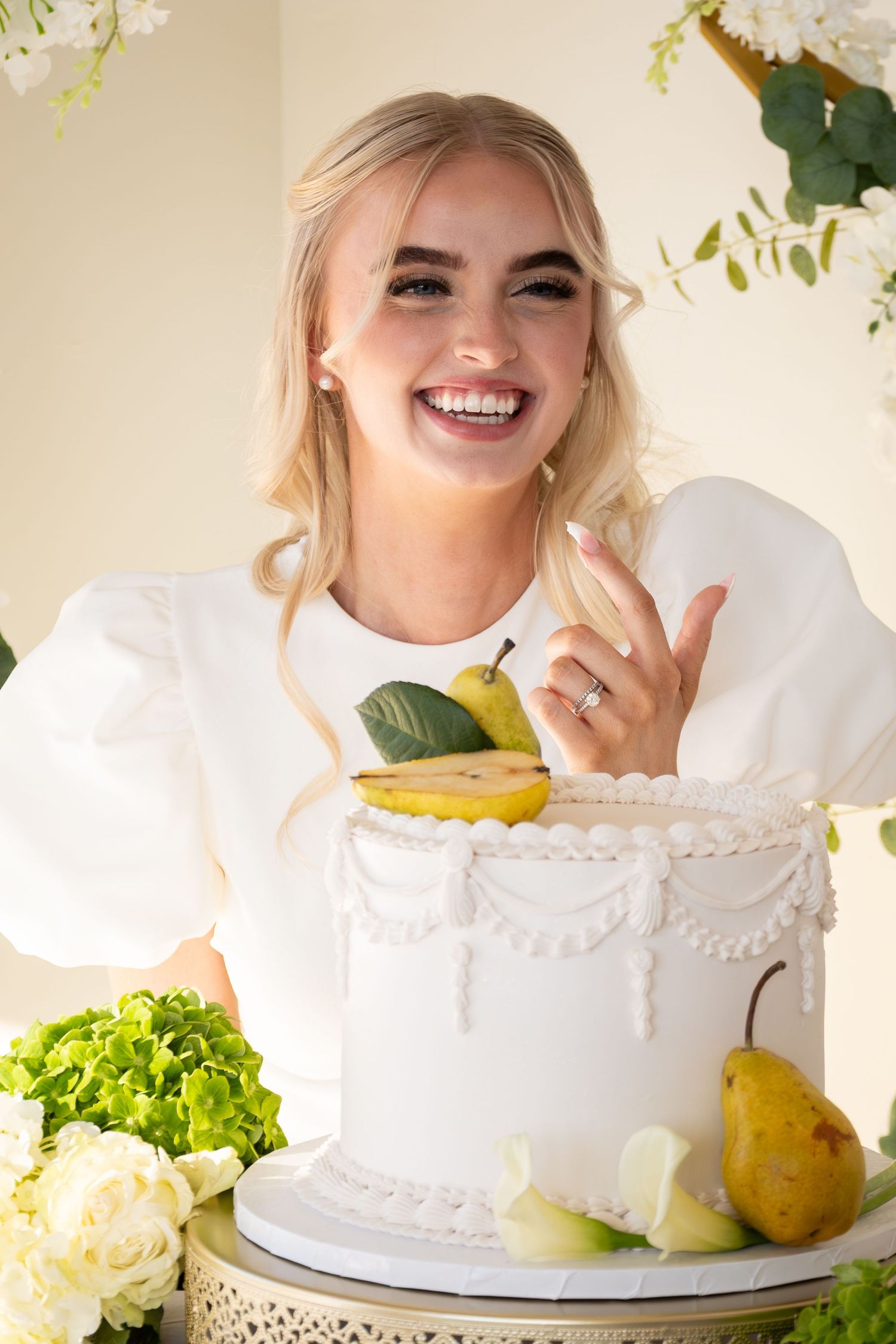 Smiling blonde bride in white dress beside elegant white wedding cake decorated with pears.
