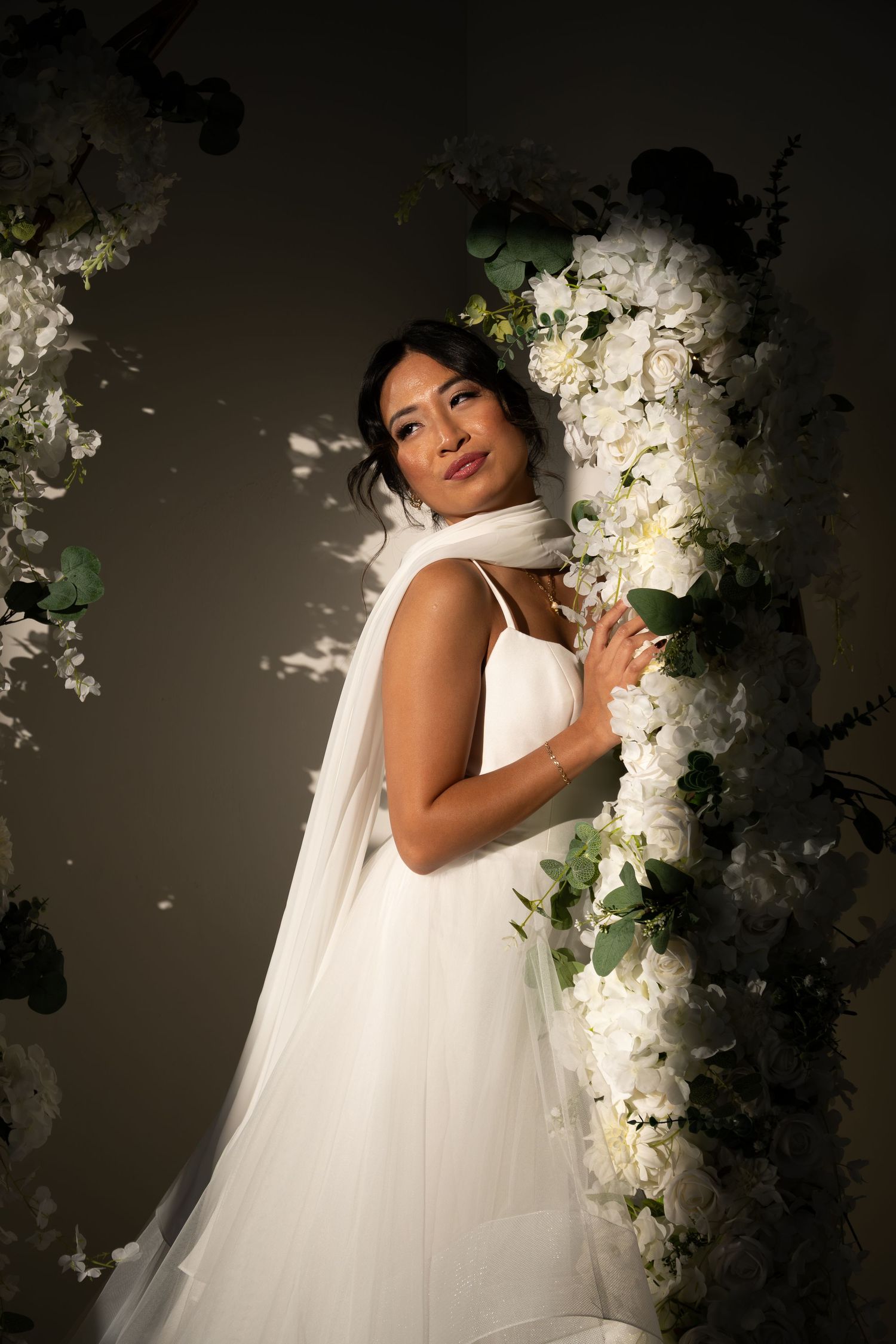 Elegant bride in white gown posing beside floral arch with white roses and greenery.