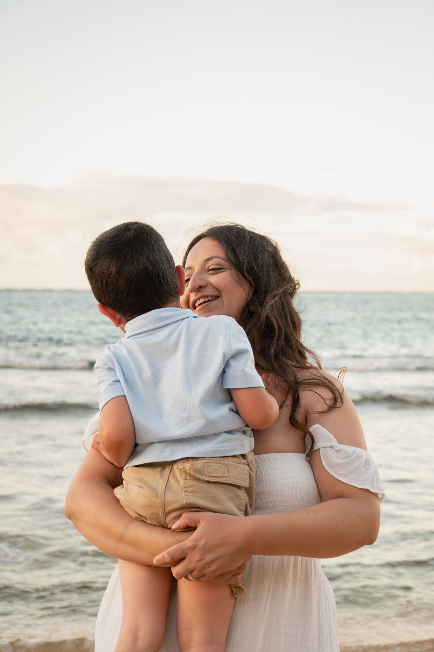 A smiling woman in a white dress holds a young boy on a beach at sunset, sharing a joyful embrace.