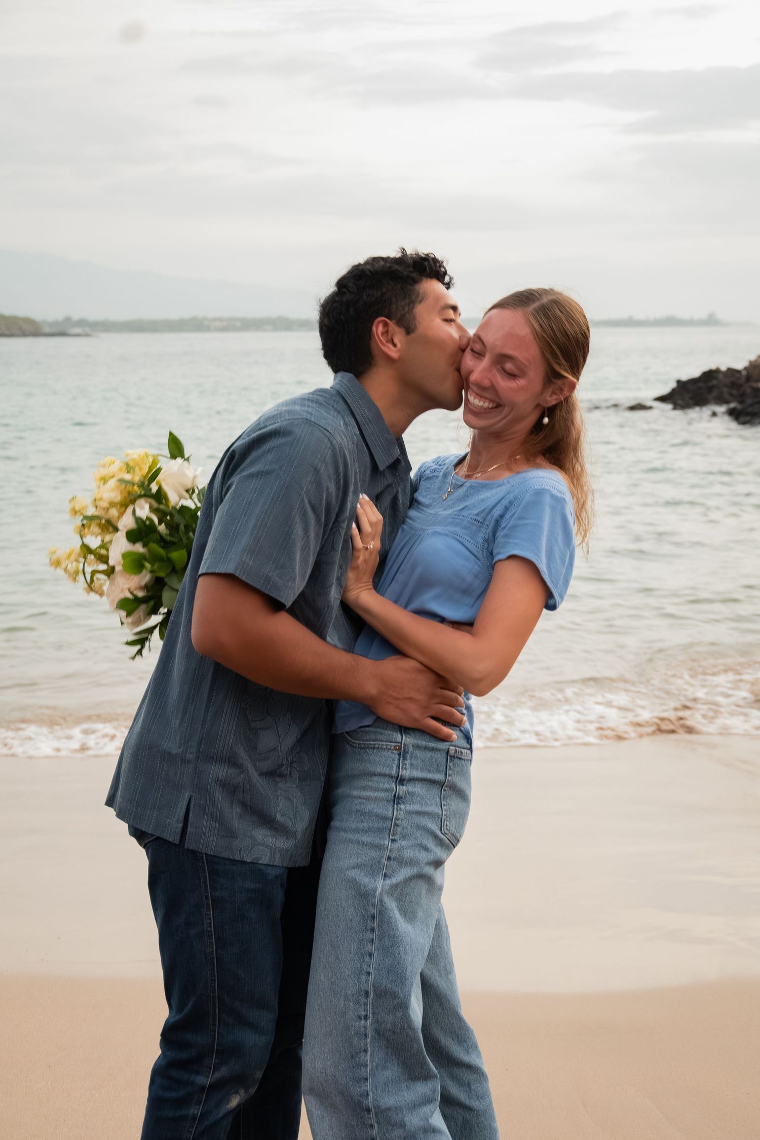 Couple sharing a romantic kiss on a beach, man holding white flowers, both wearing blue outfits with ocean backdrop.