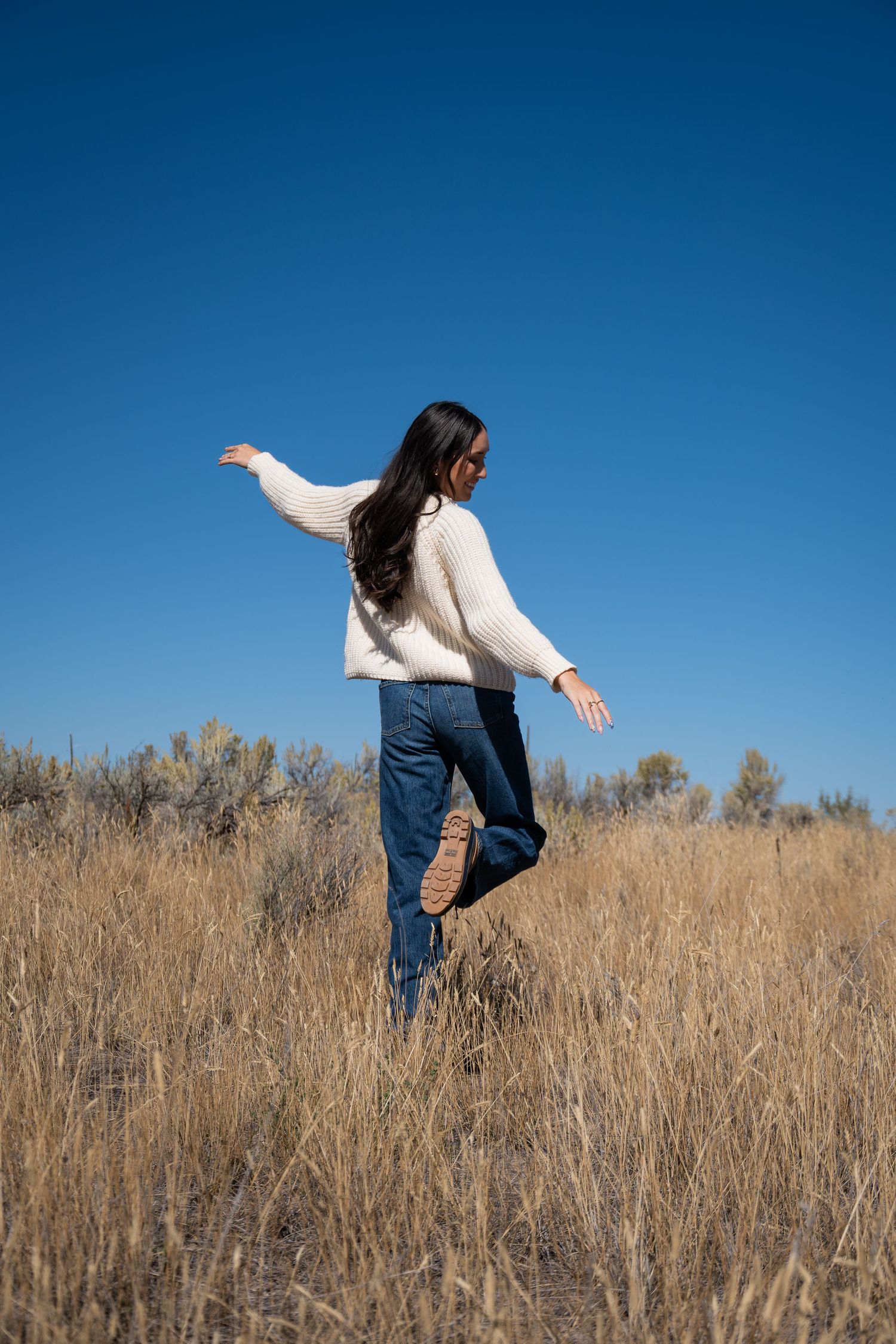 A person in a white sweater and jeans balances gracefully in a field of tall dried grass against a bright blue sky.