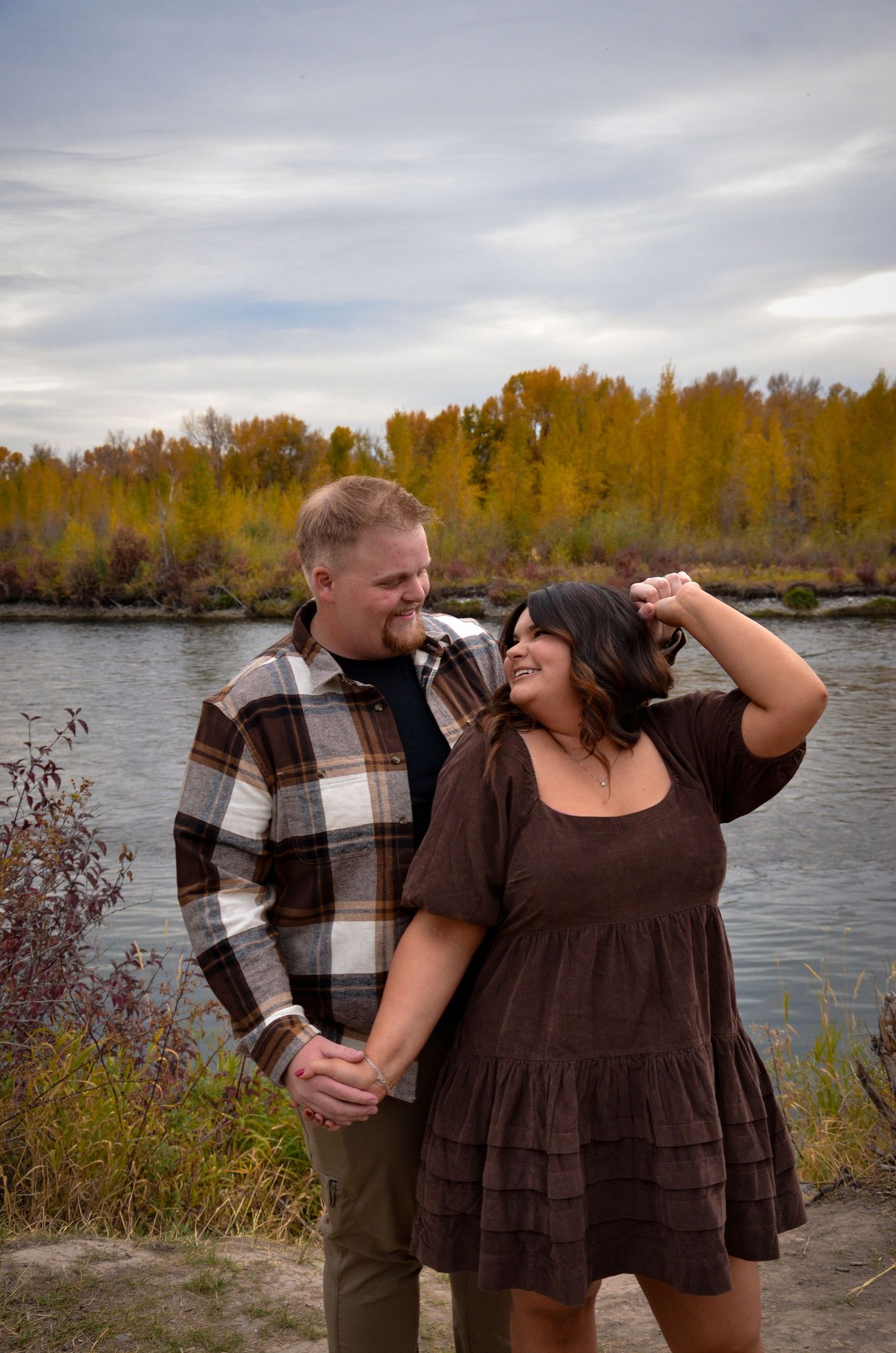 Couple sharing a playful moment by a river with autumn trees and mountains in the background.