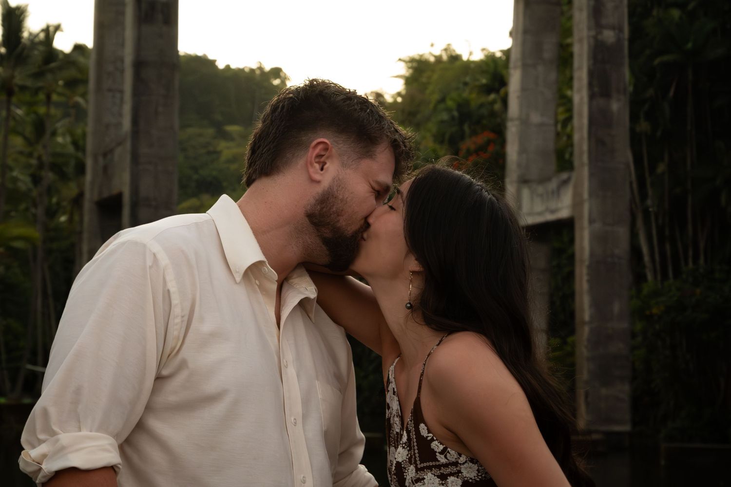 A romantic couple in white attire share an intimate kiss in an outdoor garden setting at sunset.