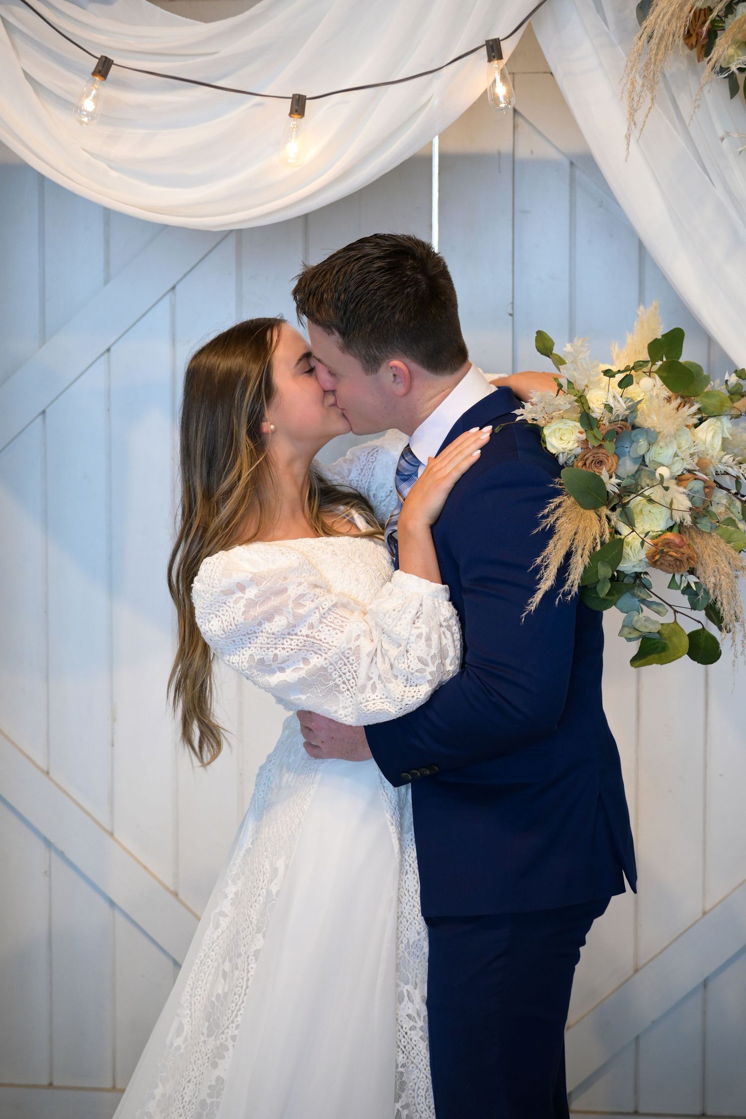 Newlyweds share a romantic kiss under white drapery and string lights at their wedding ceremony.