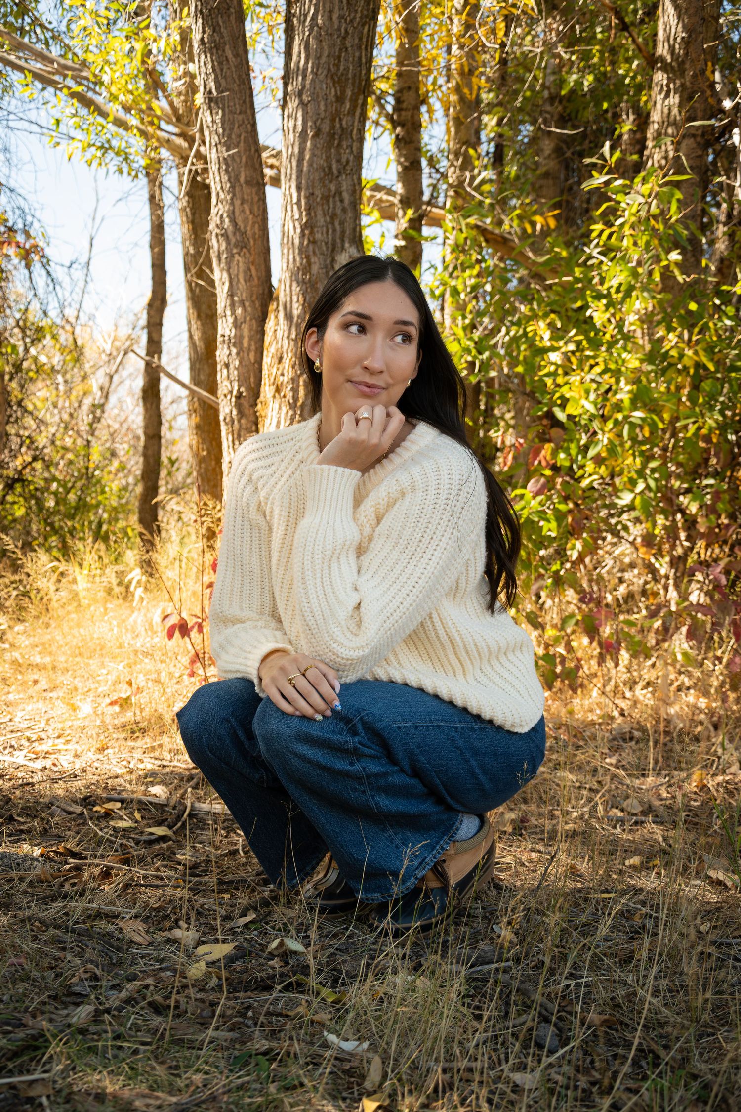 Person wearing a white knit sweater and jeans sits casually on a hillside surrounded by autumn trees and golden light.