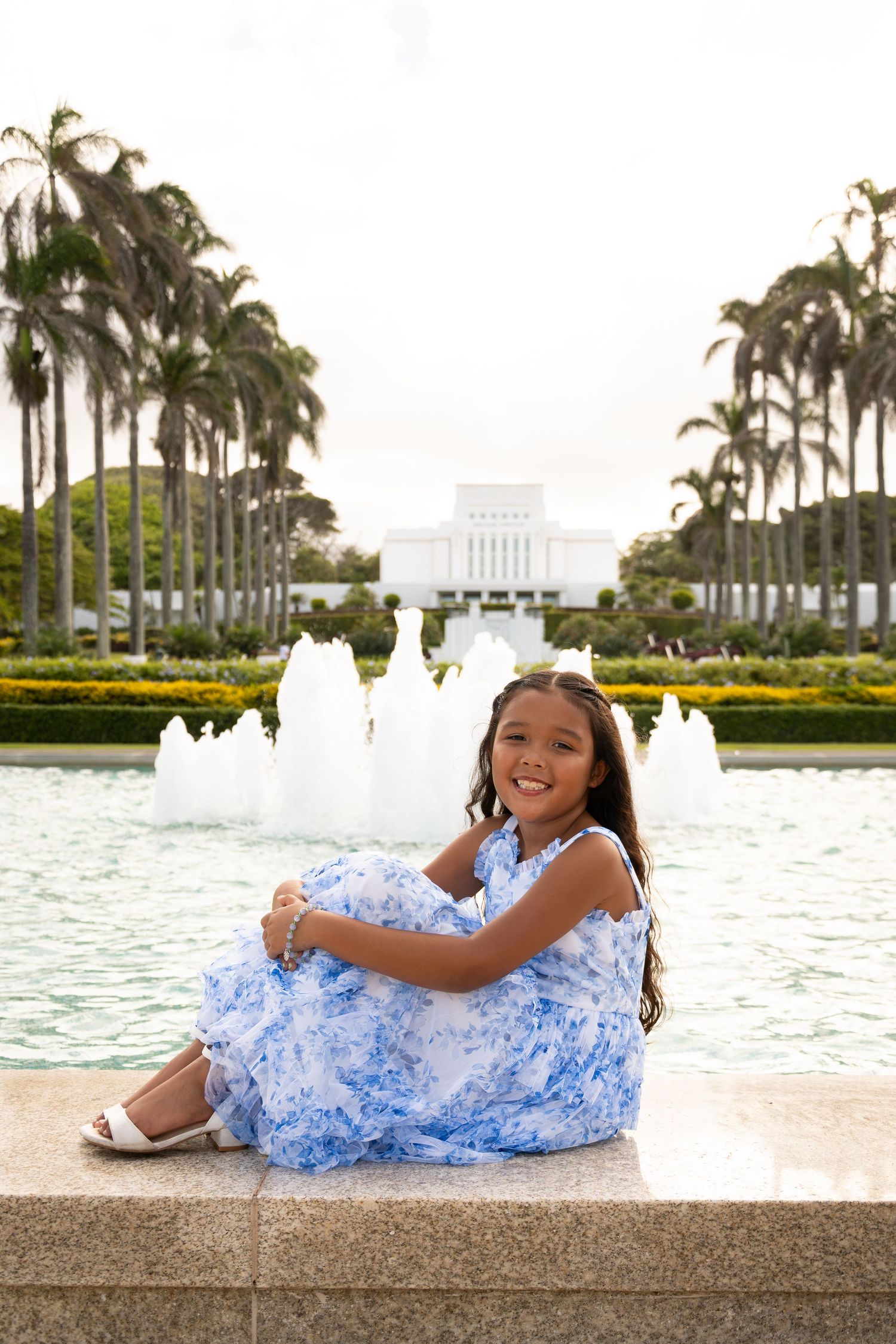 Young girl in blue floral dress sitting by fountain with palm trees and white temple building in background.