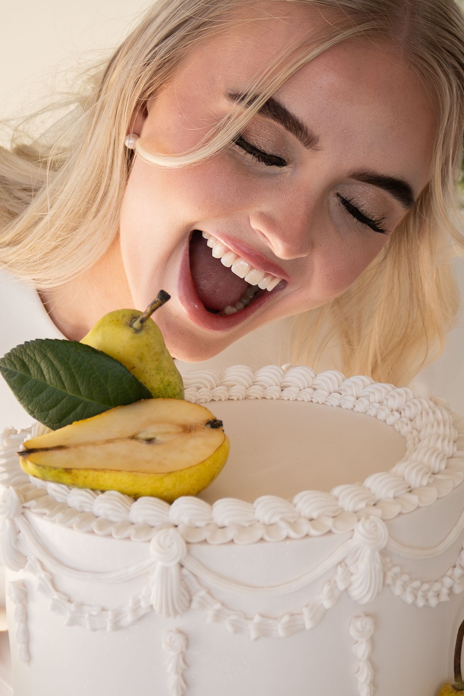 Blonde woman laughing while leaning over a white decorated cake topped with fresh pears and green leaves.