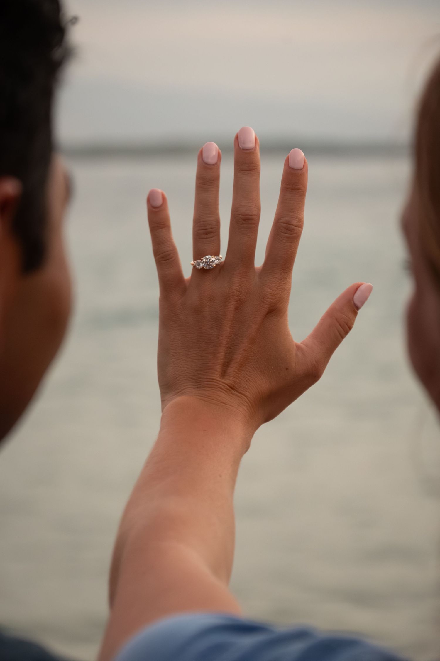 Woman displaying diamond engagement ring on ring finger with ocean backdrop, pink manicured nails, romantic proposal moment.