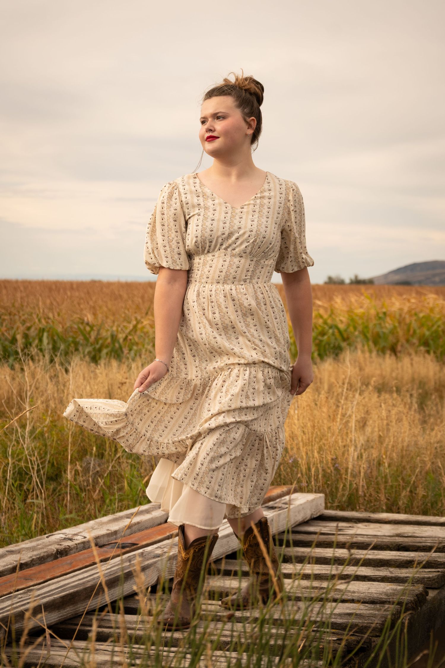 Woman in floral prairie dress and brown boots standing on rustic wooden structure in golden field.