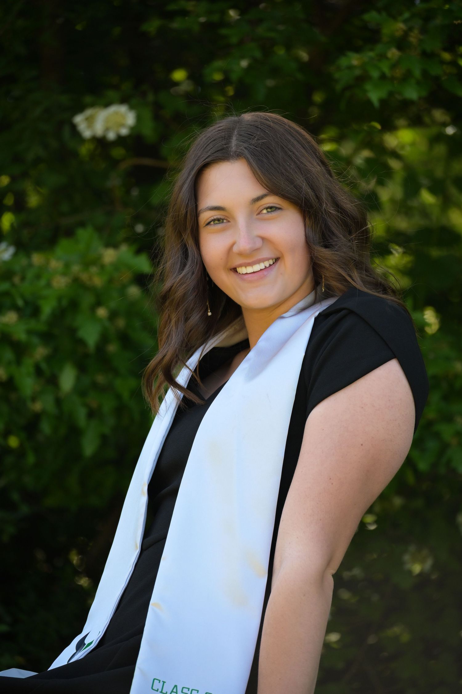 A graduate in a black cap and gown poses outdoors against a natural green background with trees.
