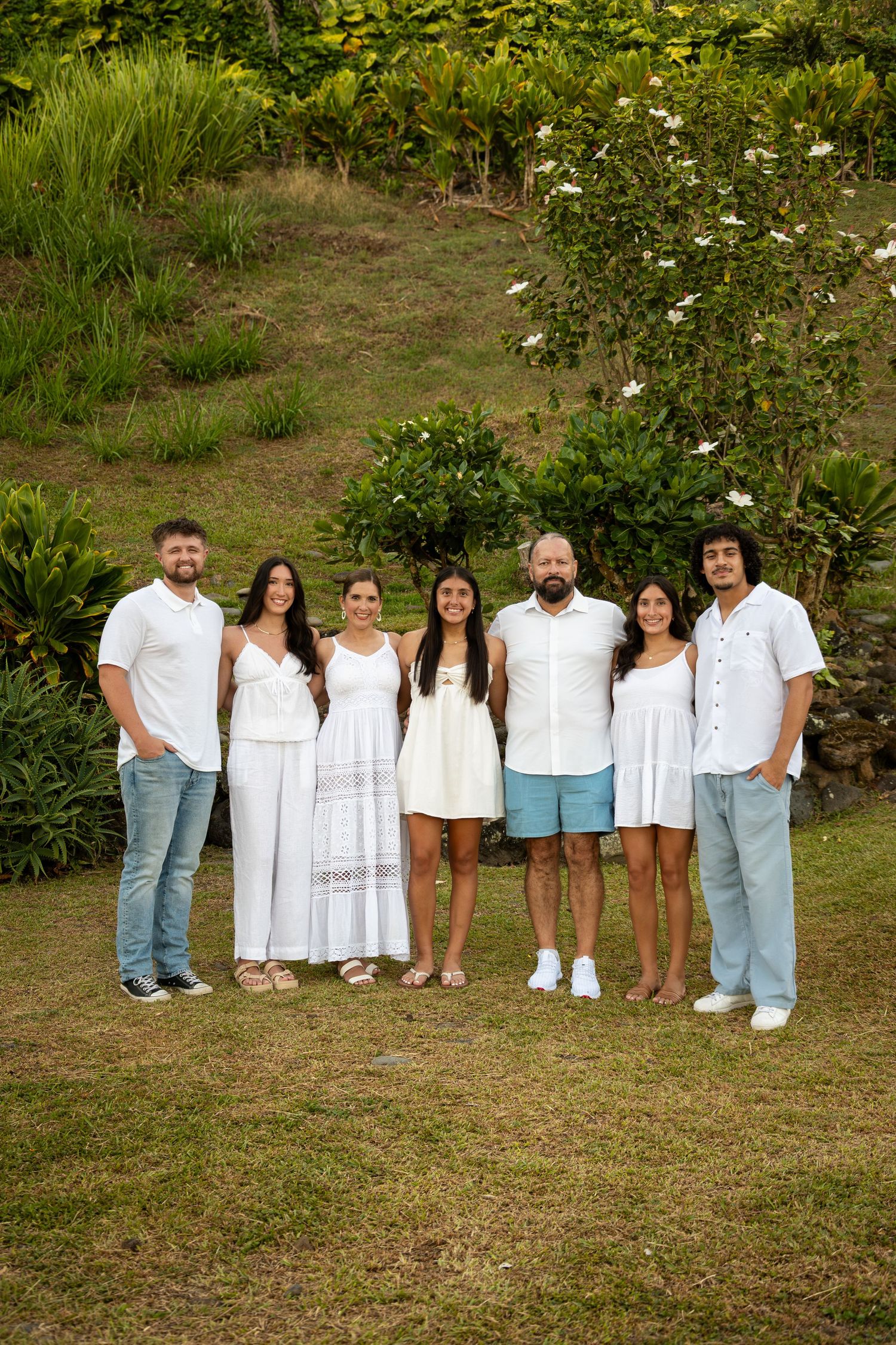 A group in coordinated white outfits poses together on a grassy hill with lush greenery in the background.