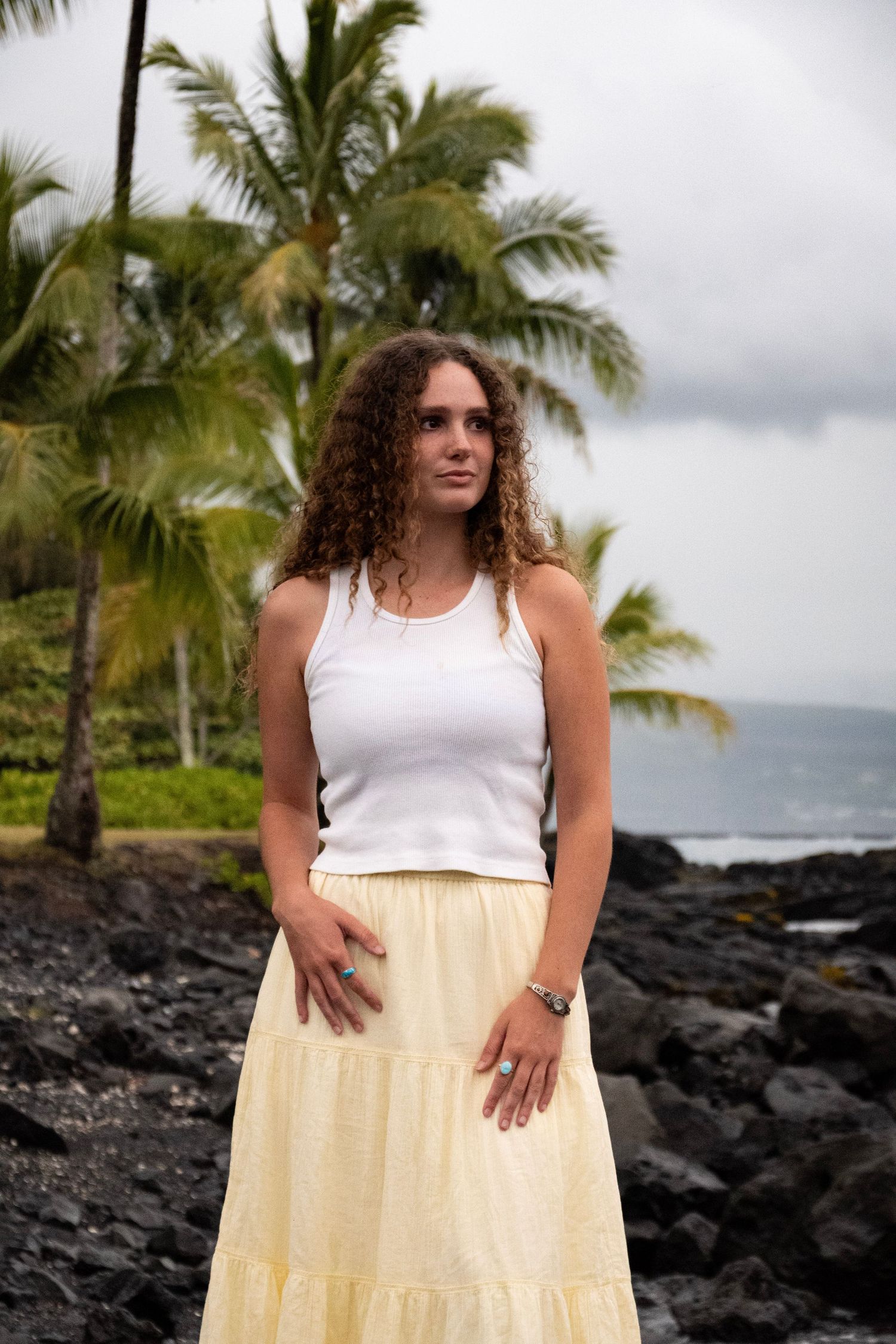 A person in a white top and yellow skirt poses on a black lava rock beach with palm trees in Hawaii.