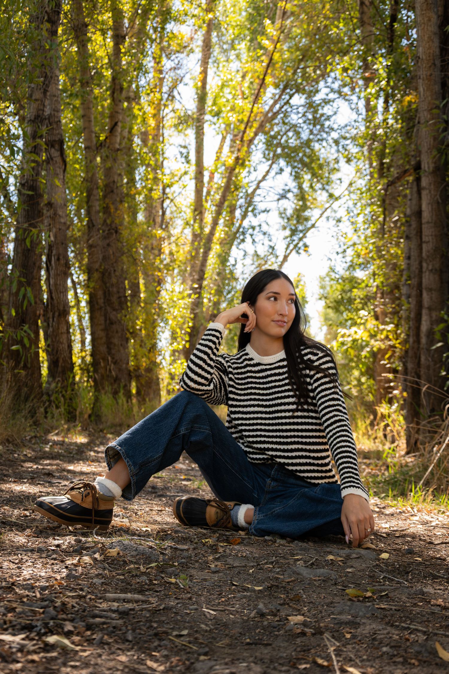 Someone wearing a striped sweater and jeans sits casually on a forest path surrounded by autumn trees in golden light.