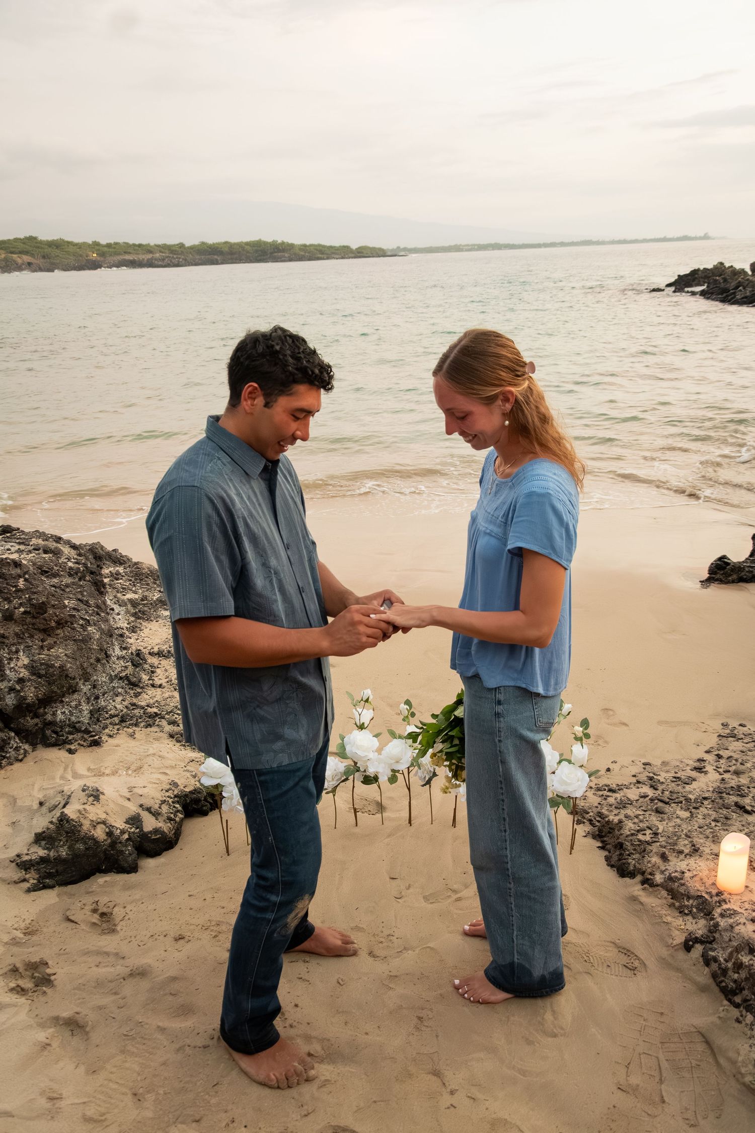 A man places a ring on a woman's finger during a romantic beach proposal at sunset, with white roses and candles in the sand.