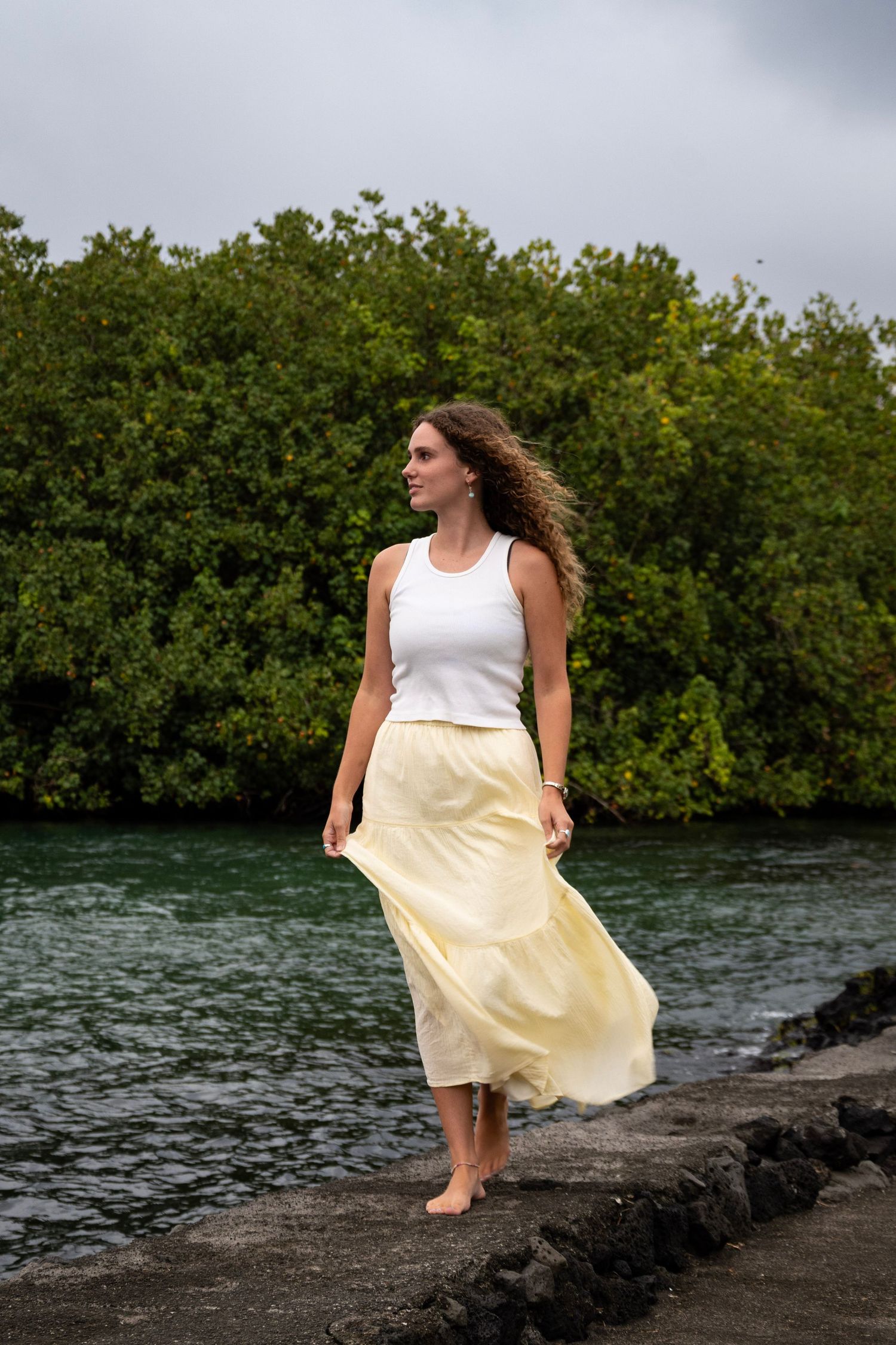 A woman in a white top and yellow skirt sits gracefully on dark rocks by the ocean with mangroves in the background.