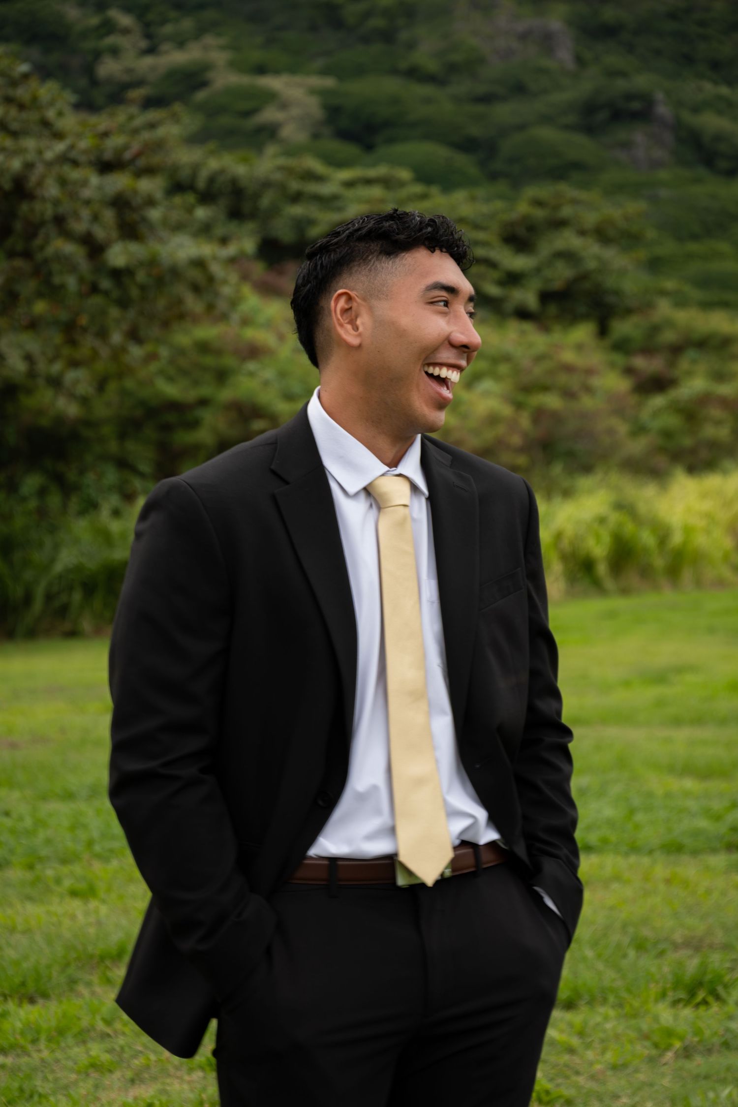 Smiling young man in black suit and gold tie standing on green grass with tropical mountains in background.