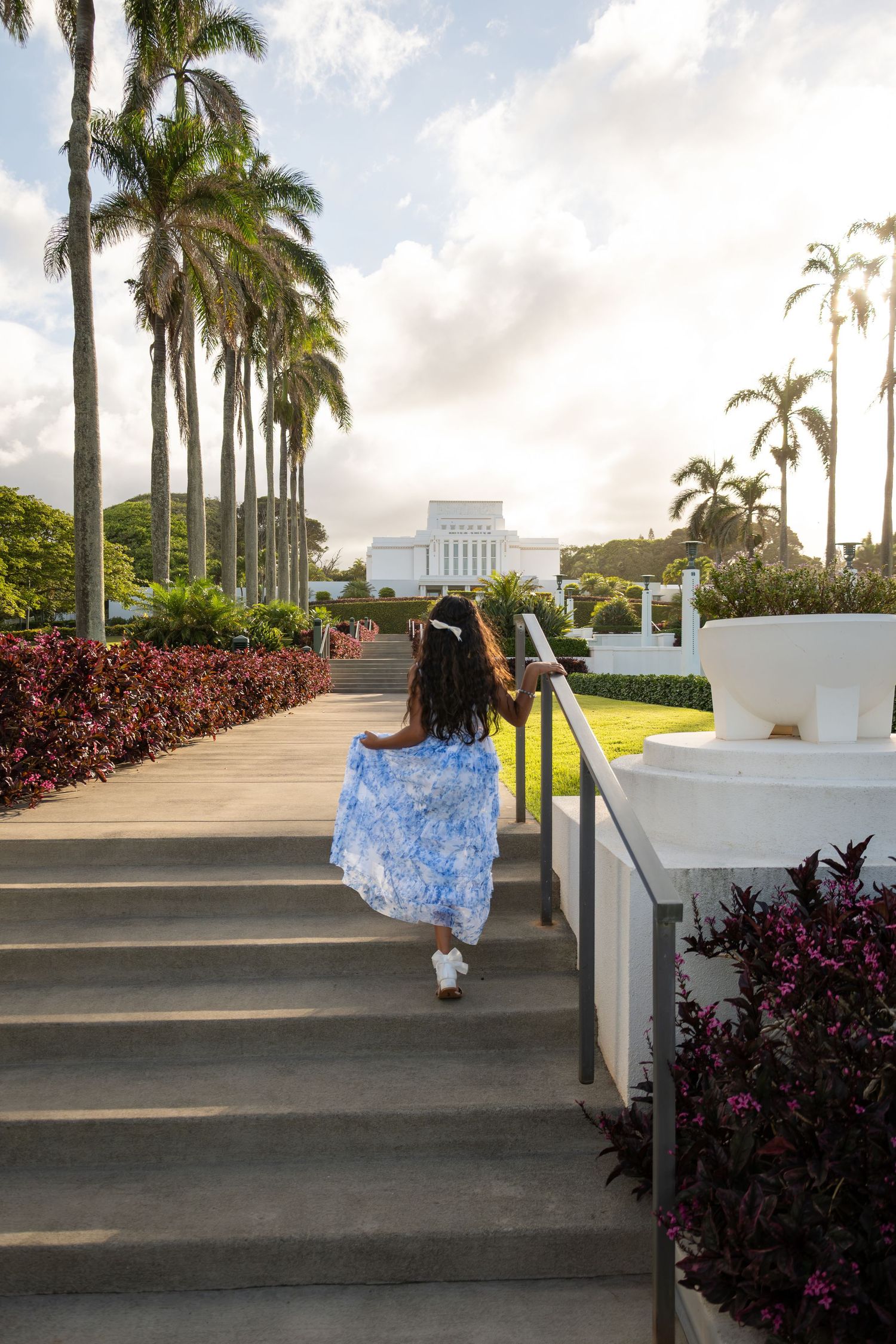 A young girl in a blue floral dress walks up steps toward a white temple surrounded by tall palm trees in Hawaii.
