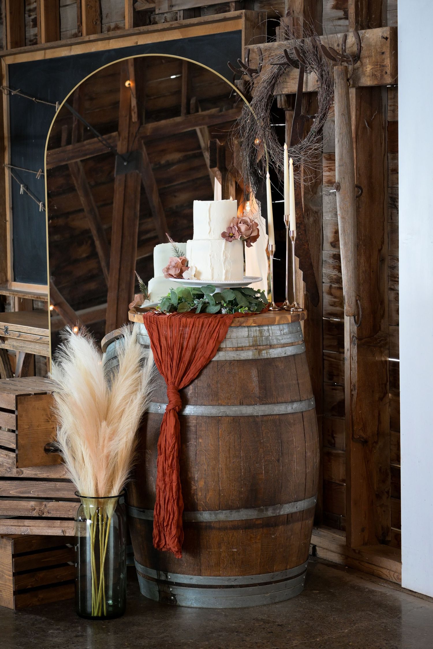 Wedding cake display on wooden barrel with pampas grass and macrame decor in rustic venue.