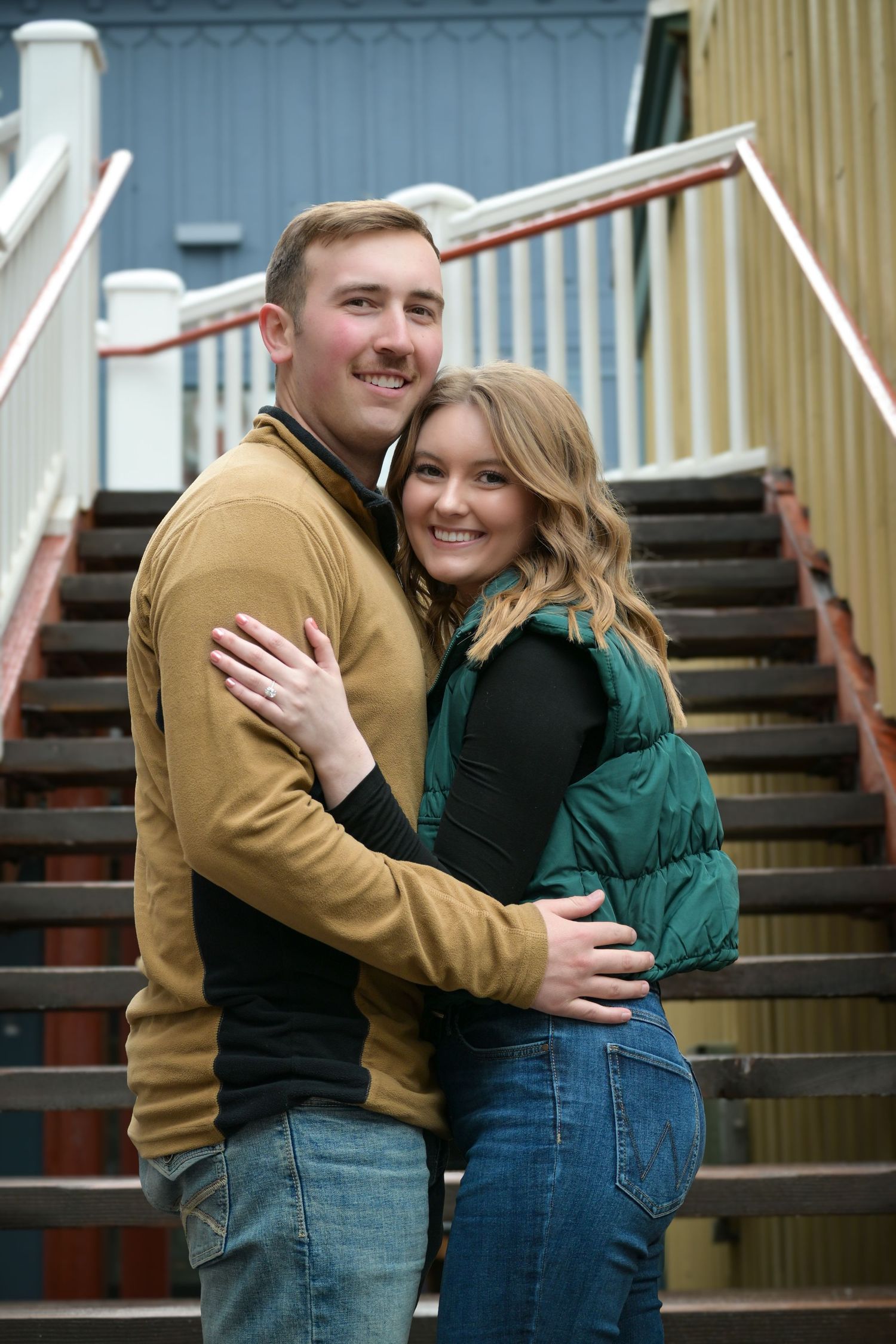 A couple embraces on outdoor stairs, dressed in tan and green winter layers.
