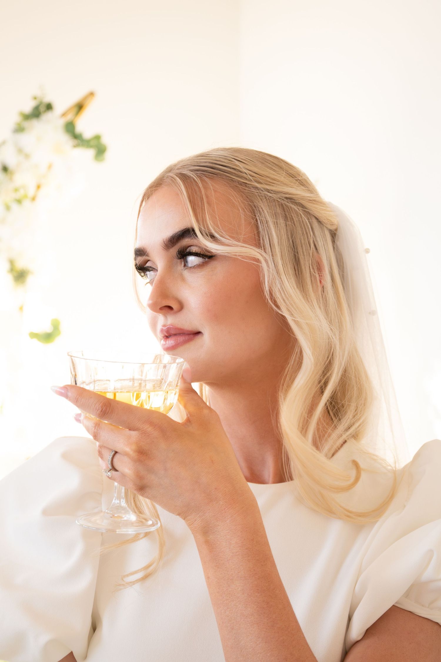 Elegant blonde bride in white dress holding a crystal glass, gazing upward in soft light.