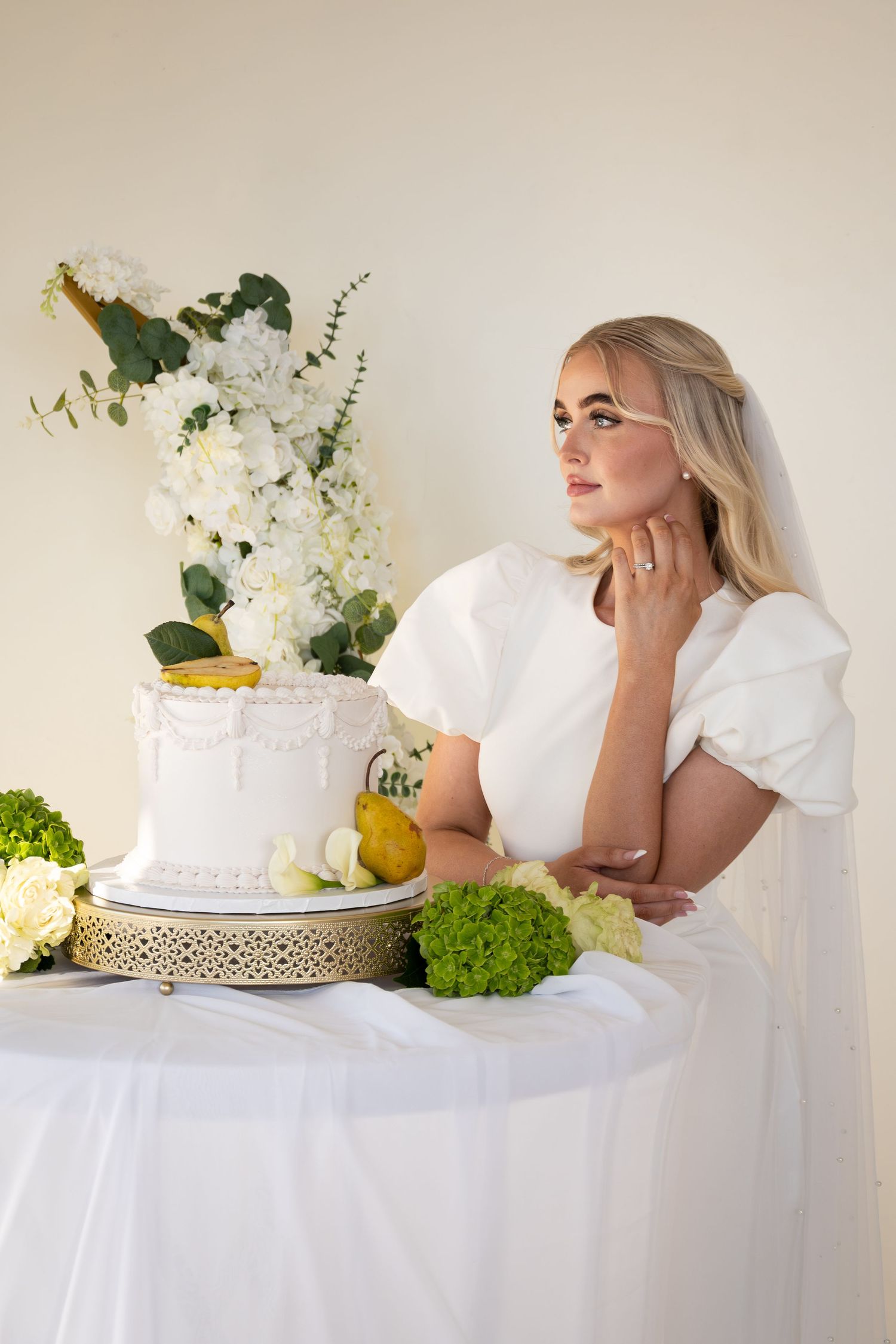 Elegant bride in white dress with puff sleeves sits beside a tiered wedding cake adorned with flowers and fruit.