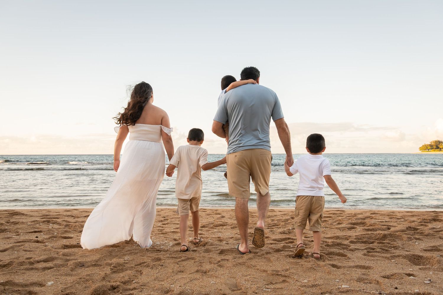 Family of four walking hand-in-hand on a sandy beach at sunset, viewed from behind near ocean waves.