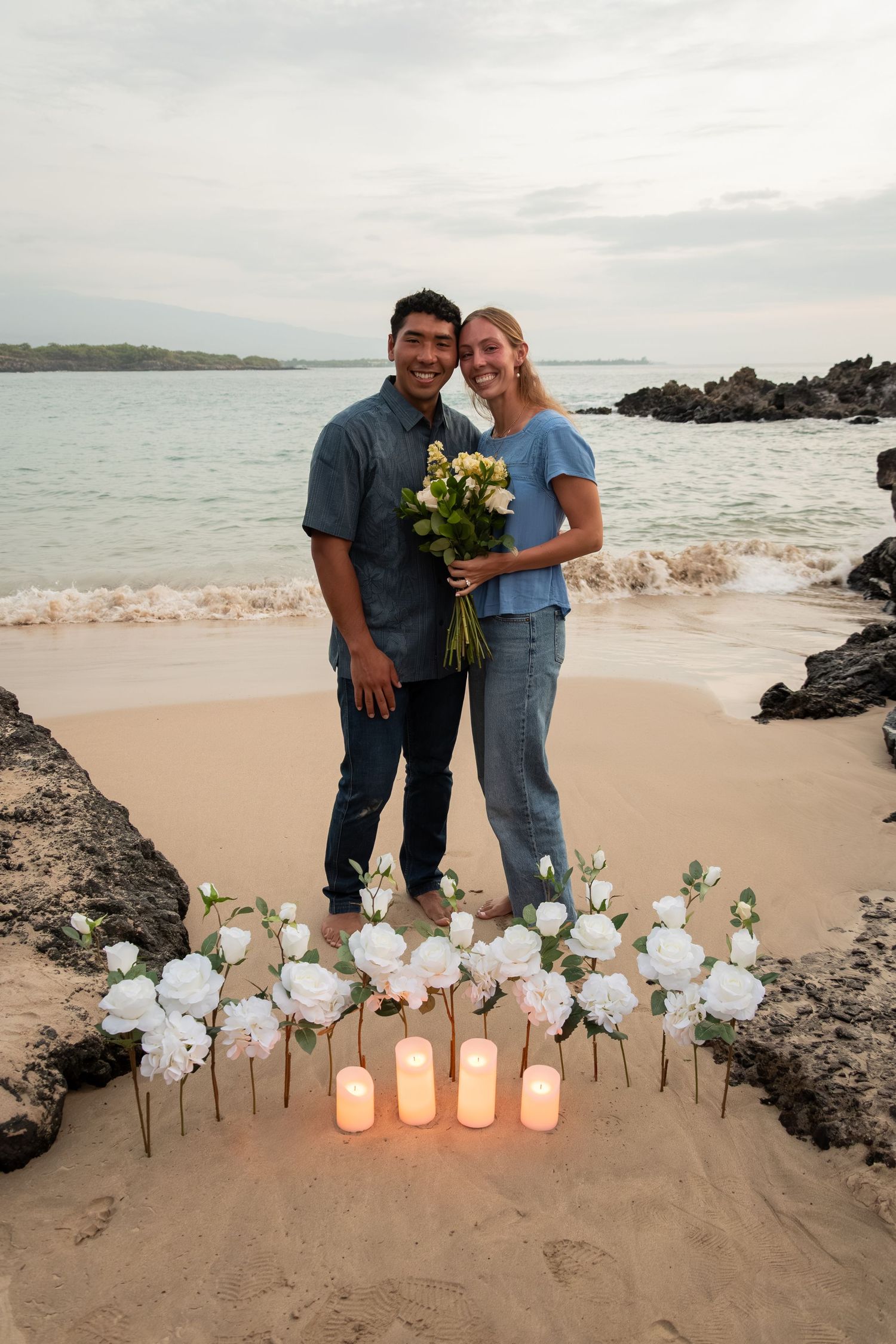 Couple smiling on a beach at sunset with white roses and candles arranged in the sand, holding a bouquet.