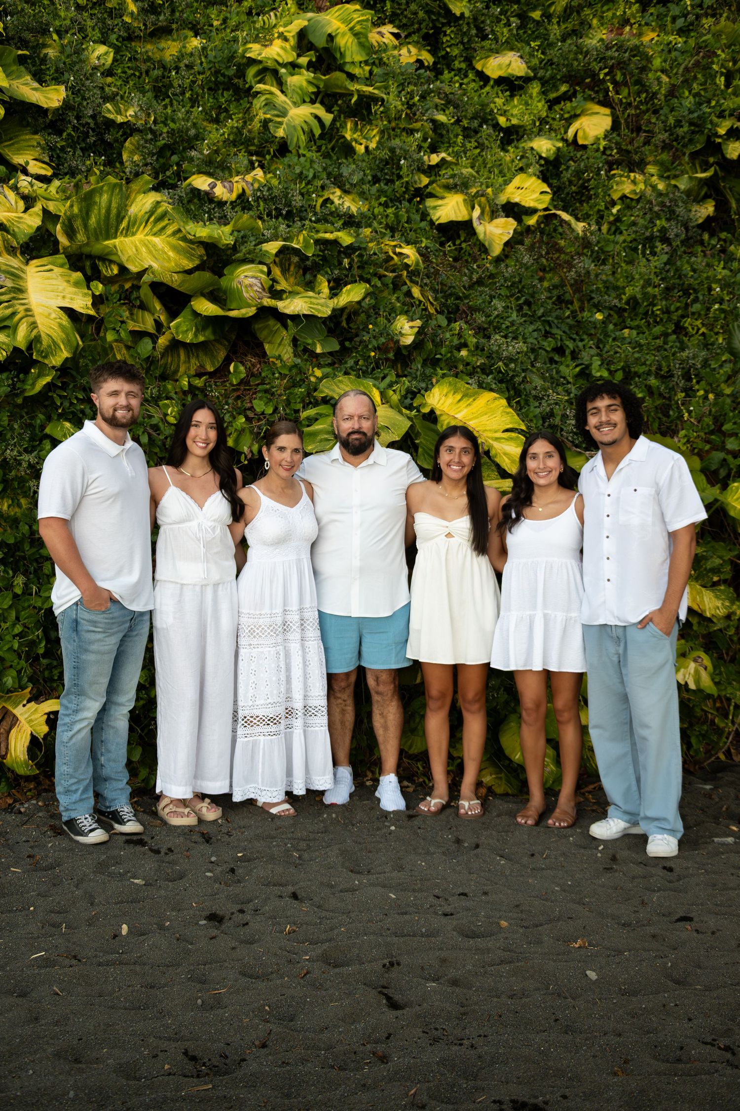 A group of people wearing white and light-colored clothing pose together on a black sand beach with tropical foliage behind them.